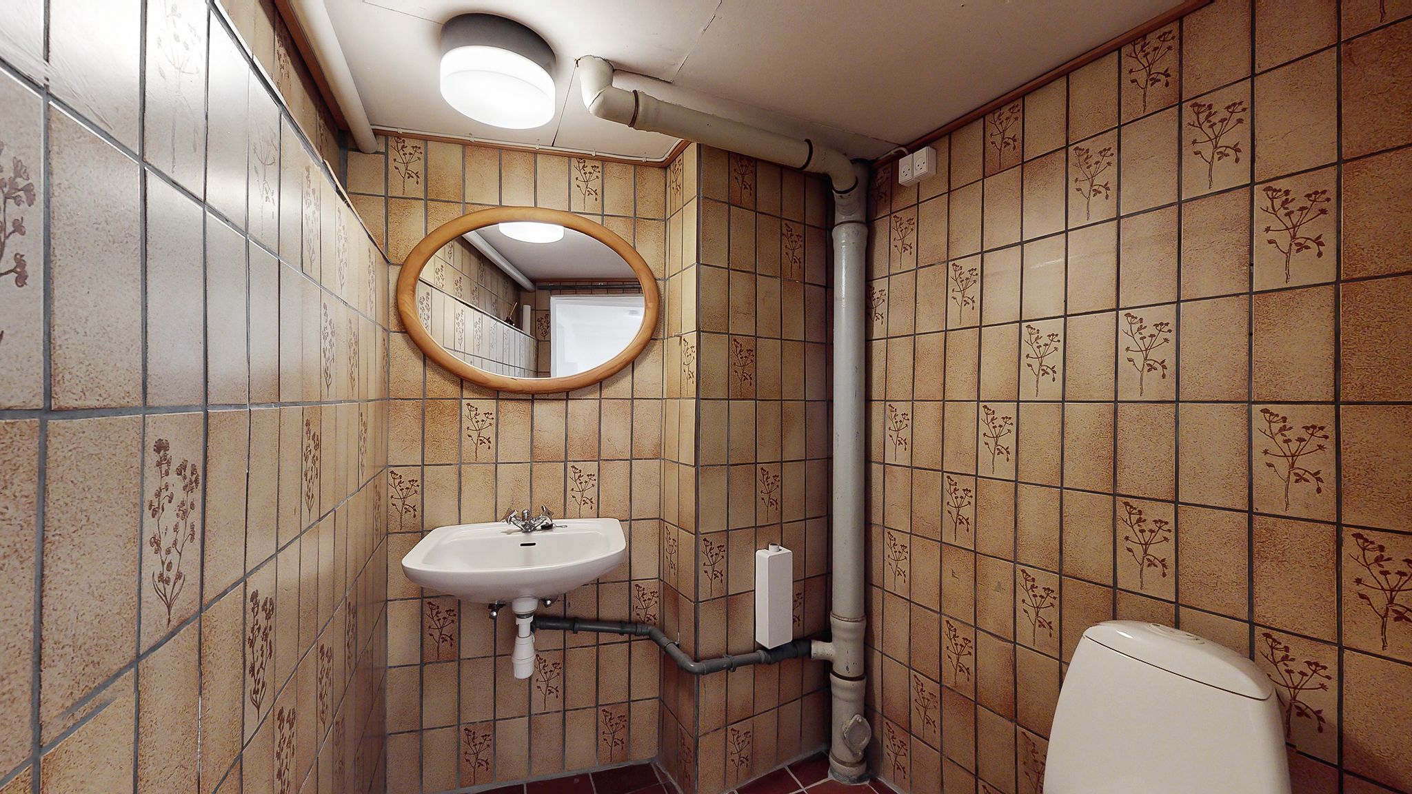 En lille badeværelse med lysegult belagte vægge og toilet i højre side. Der er et rundt spejl og en hvid vask med