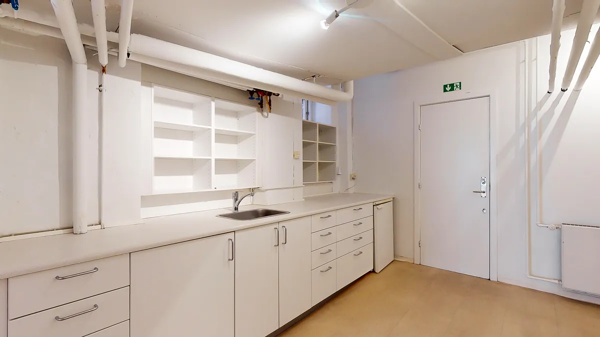 Funktionelt værelse med hvide vægge, hvide køkkenskabe og en vask med vandhane og opvaskemaskine i hjørnet