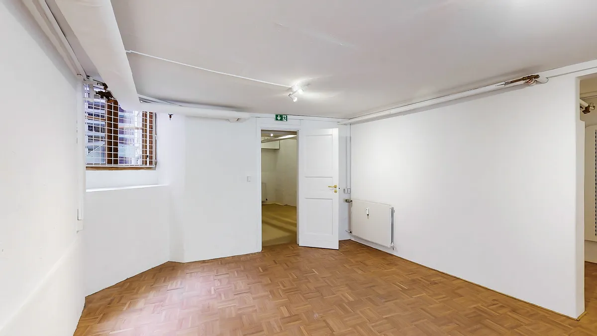 Et funktionelt butikslejemål med hvide vægge, trægulv og et slankt, hvidt dørblad til siden