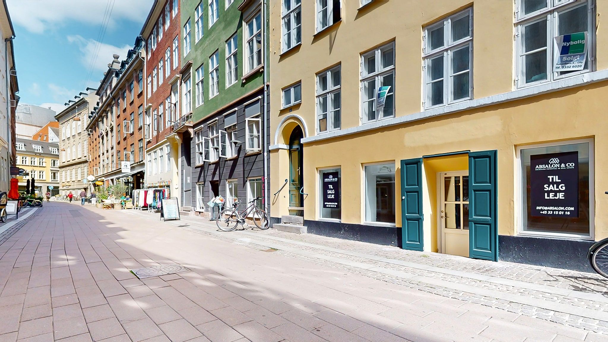 Lejlighedsgade med butikker og sammenlagte cykler foran butikken med teksten "TIL SALG LEJE" på døren.
