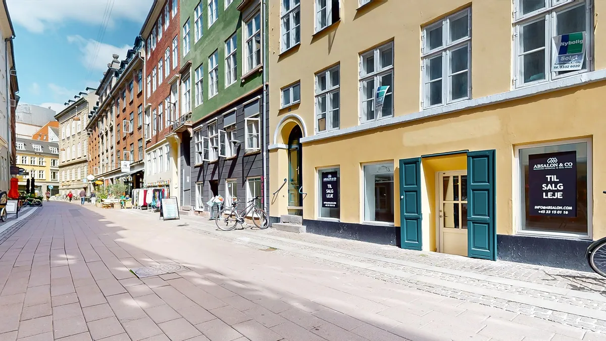Lejlighedsgade med butikker og sammenlagte cykler foran butikken med teksten "TIL SALG LEJE" på døren.