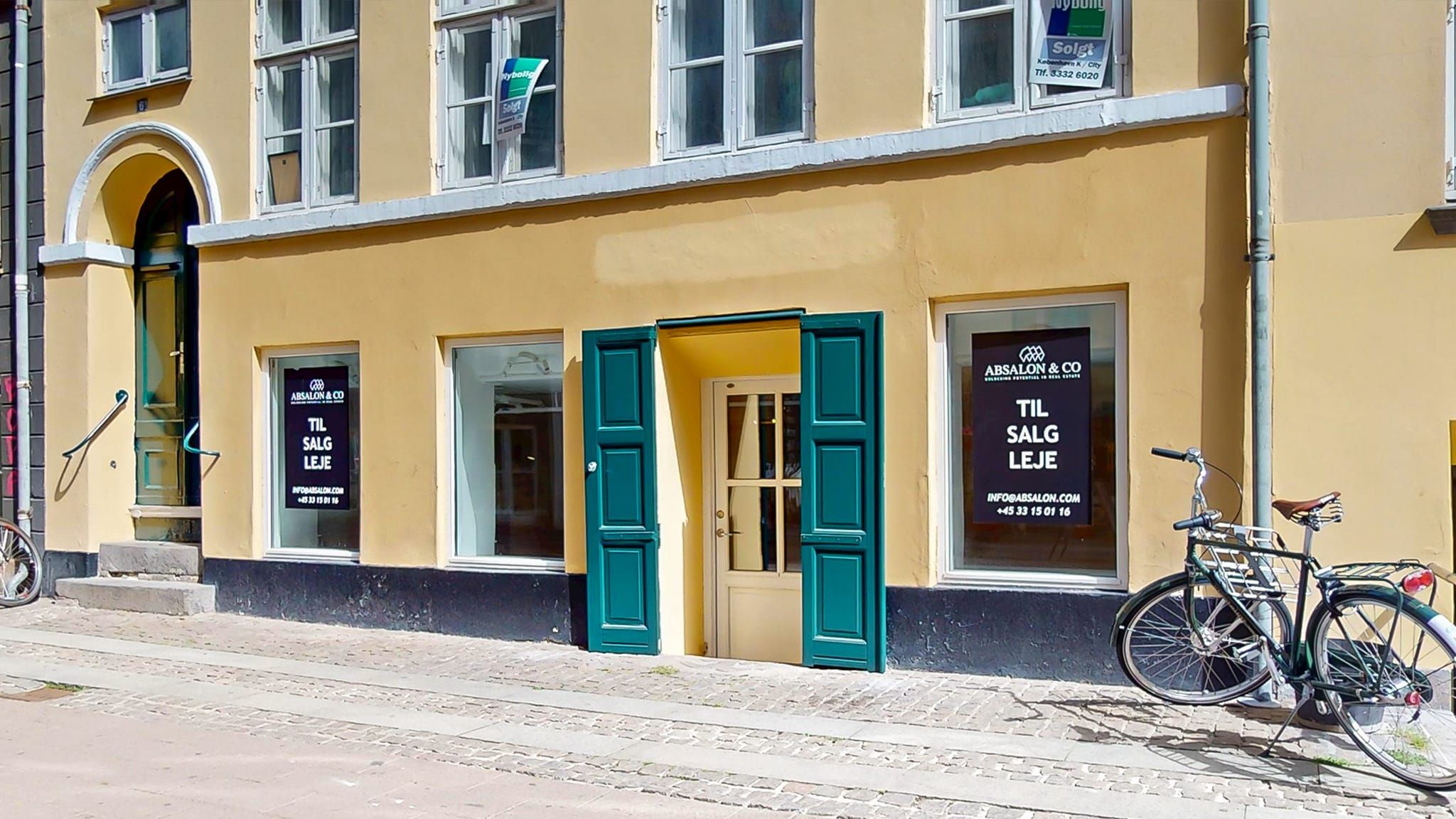 En bycykel parkeret foran en gul bygning med flere butikker og reklameskilte for forskellige lejemål