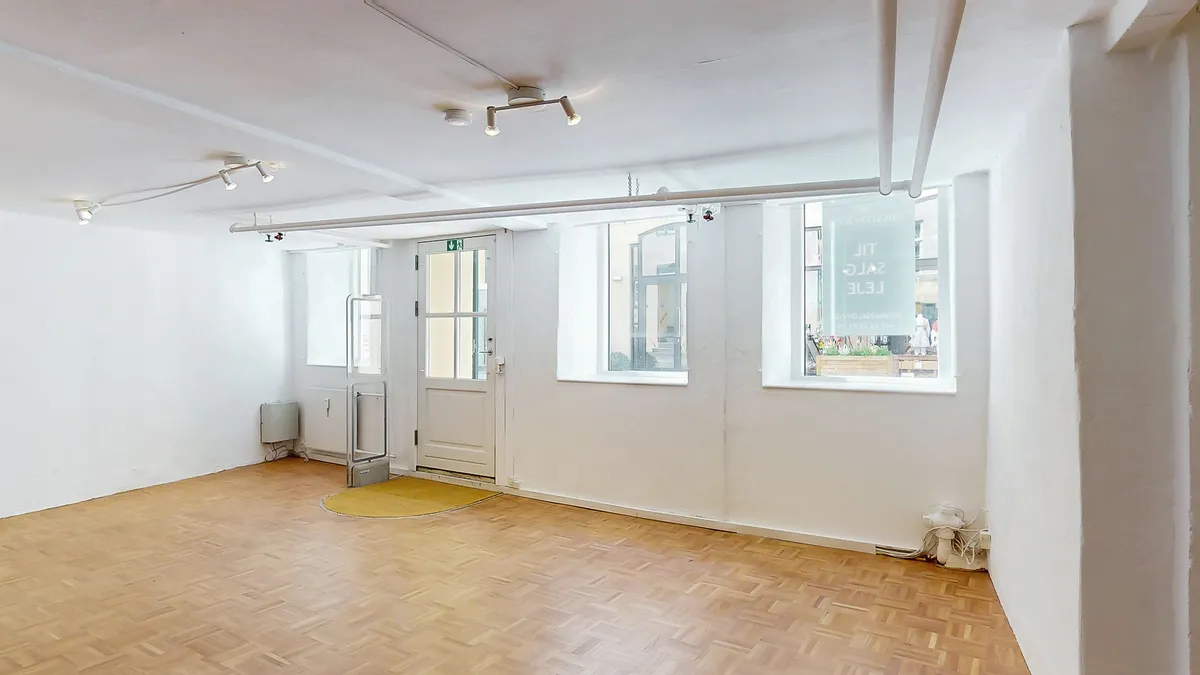 Et lejlighedslejemål med en stor åben stue, to vinduer og en hvid dør i midten af rummet