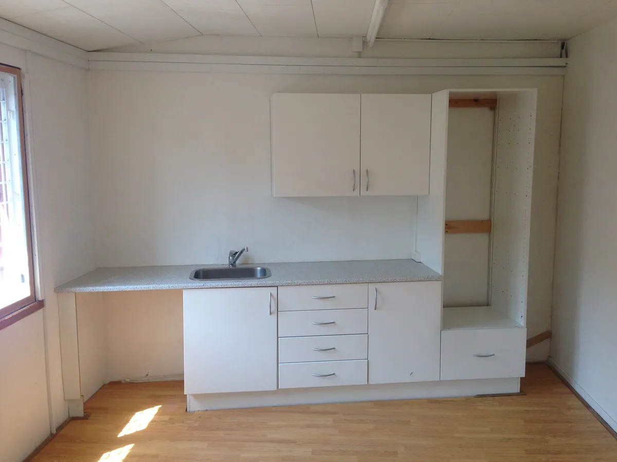 Køkken med hvide vægge og gulv samt højt tællebord og sænket vask med vandhane og højt skab med to døre