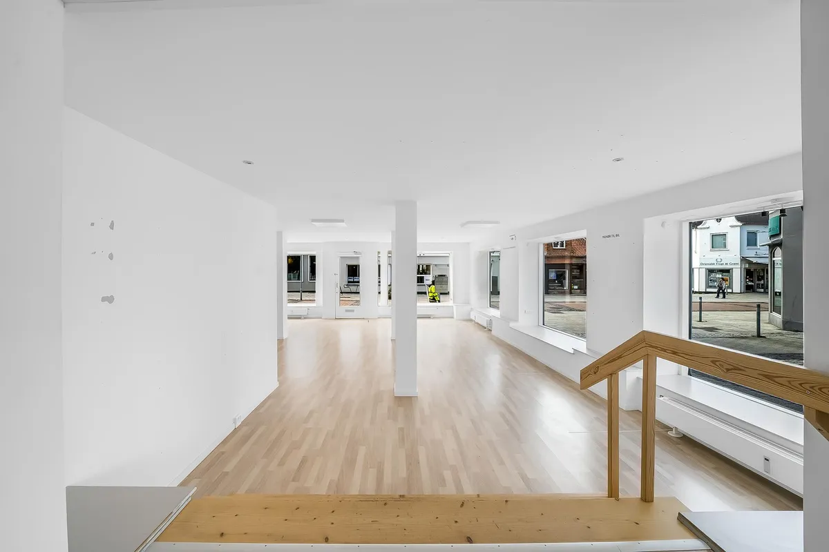 En tom butik med hvide vægge og gulv samt vinduer og trapper i højre side.