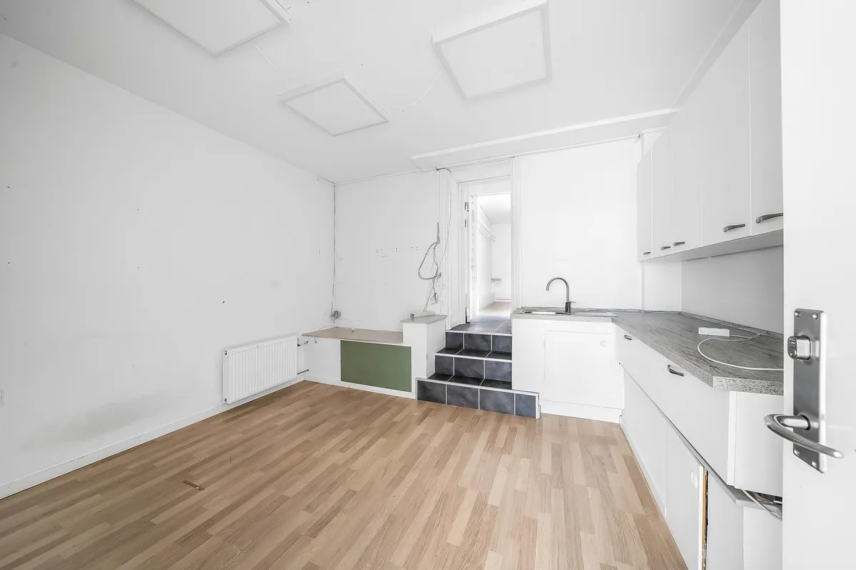 Det er et minimalistisk køkken med hvide vægge, højloft, lyse trægulve, og et lysegråt stenbord med en håndvask og en