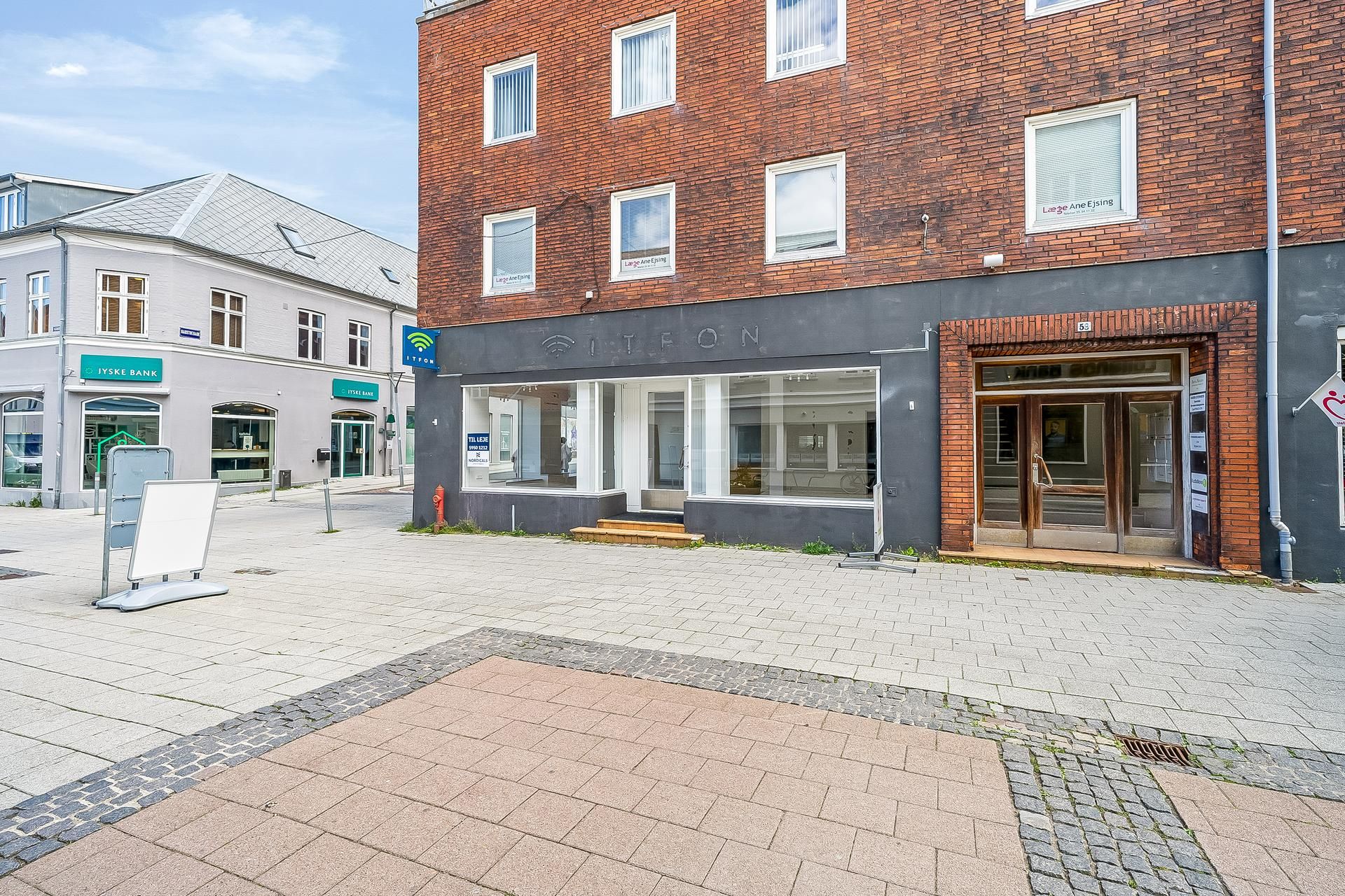 Foran en butik med en rødt murstens facade er der en forhøjning med en betonplade og et trætrin. I baggrunden ses en bygning