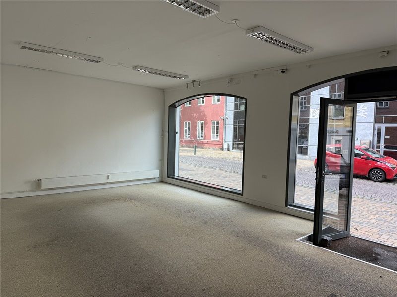 Lejlighed med store vinduer mod Ringe Centrum. Moderne, hvidt interiør med lysekroner og to døre. Rød bil parkeret udenfor.