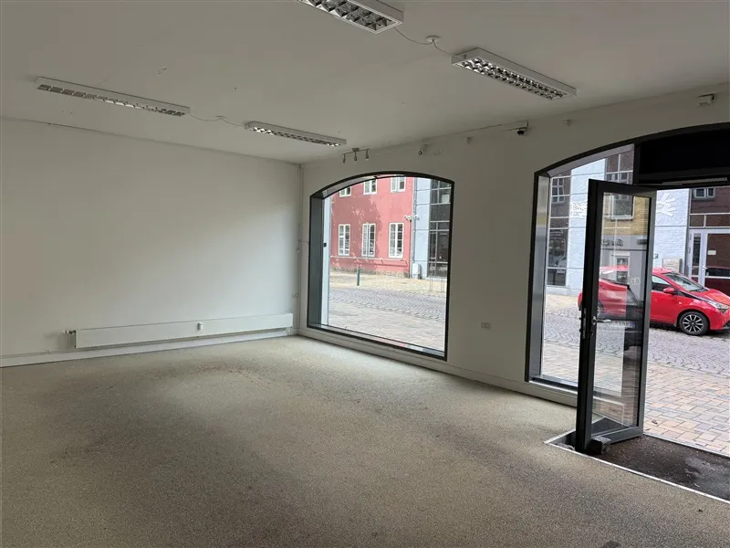 Lejlighed med store vinduer mod Ringe Centrum. Moderne, hvidt interiør med lysekroner og to døre. Rød bil parkeret udenfor.