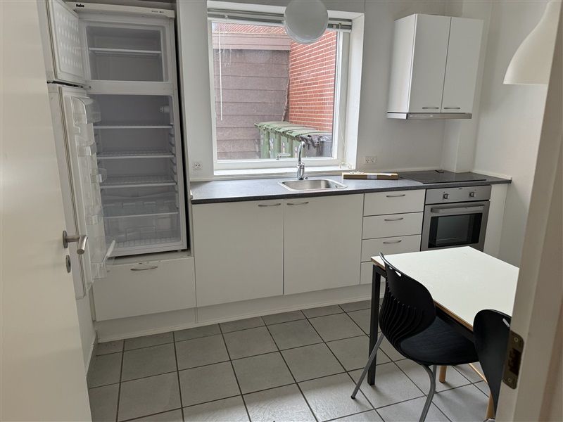 Et køkken med køleskab, komfur, bord og stole samt et vindue med udsigt til et rødmurstensbygning og skraldespande