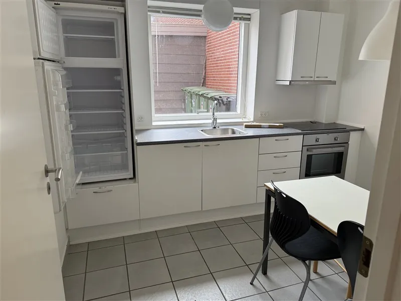 Et køkken med køleskab, komfur, bord og stole samt et vindue med udsigt til et rødmurstensbygning og skraldespande