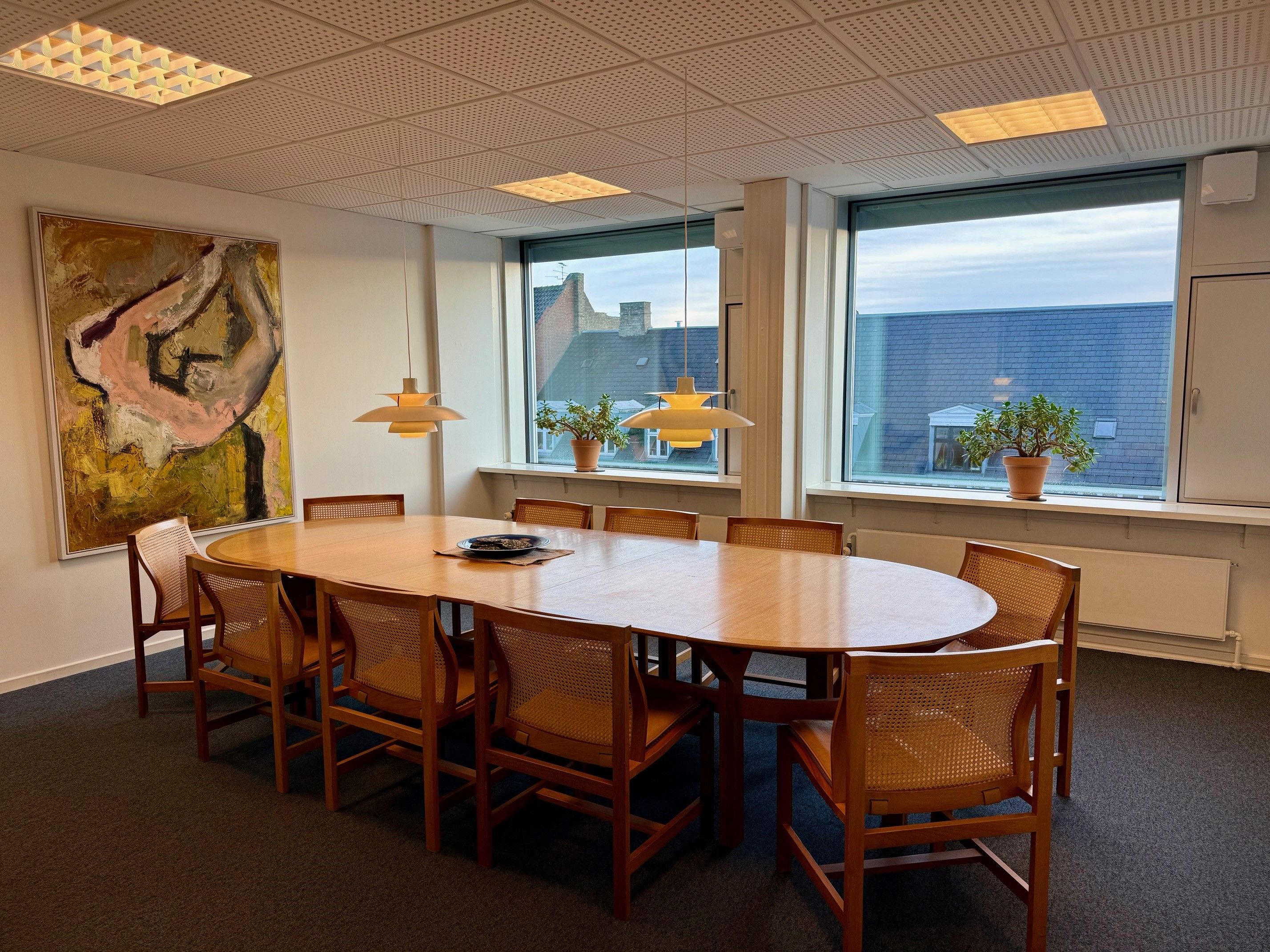 Kontor med rundt bord og stole. Planter i vindueskarmen og loftslamper. Maleri på væggen og en dør til højre.