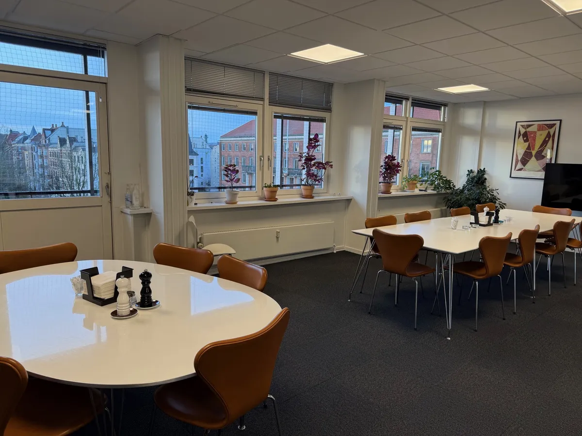 Kontorlokale med runde borde og lædersæder. En stor tv-skærm og flere blomster på vindueskarmen. Lys og ren indretning.