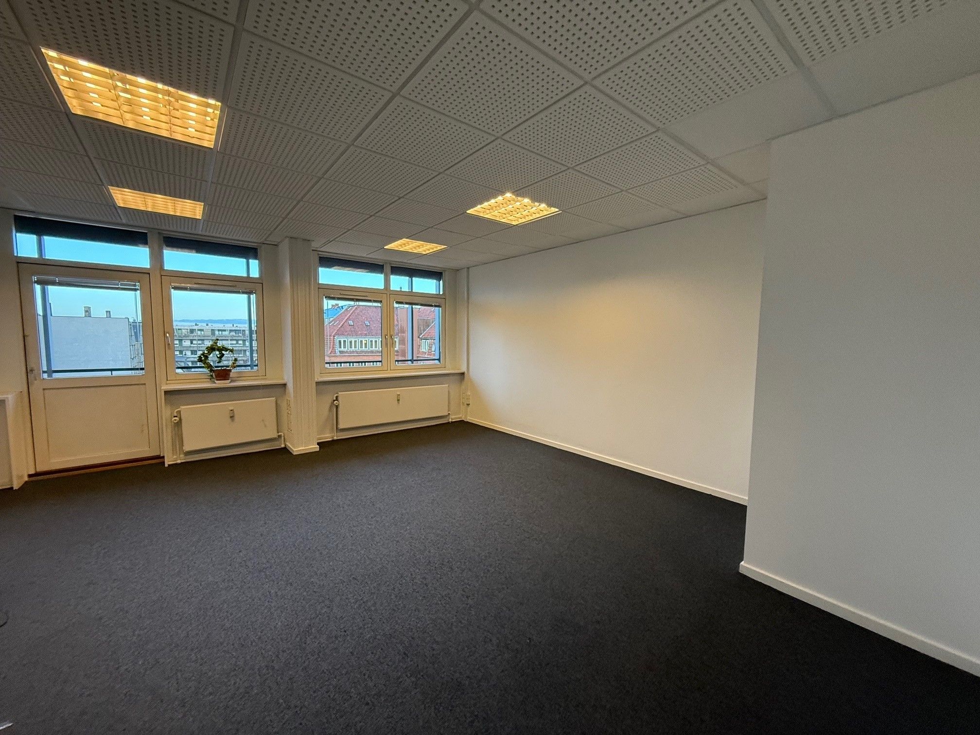 Kontorlokale med hvide vægge og sort gulv, lysekroner, tre vinduer og radiatorer under vinduerne