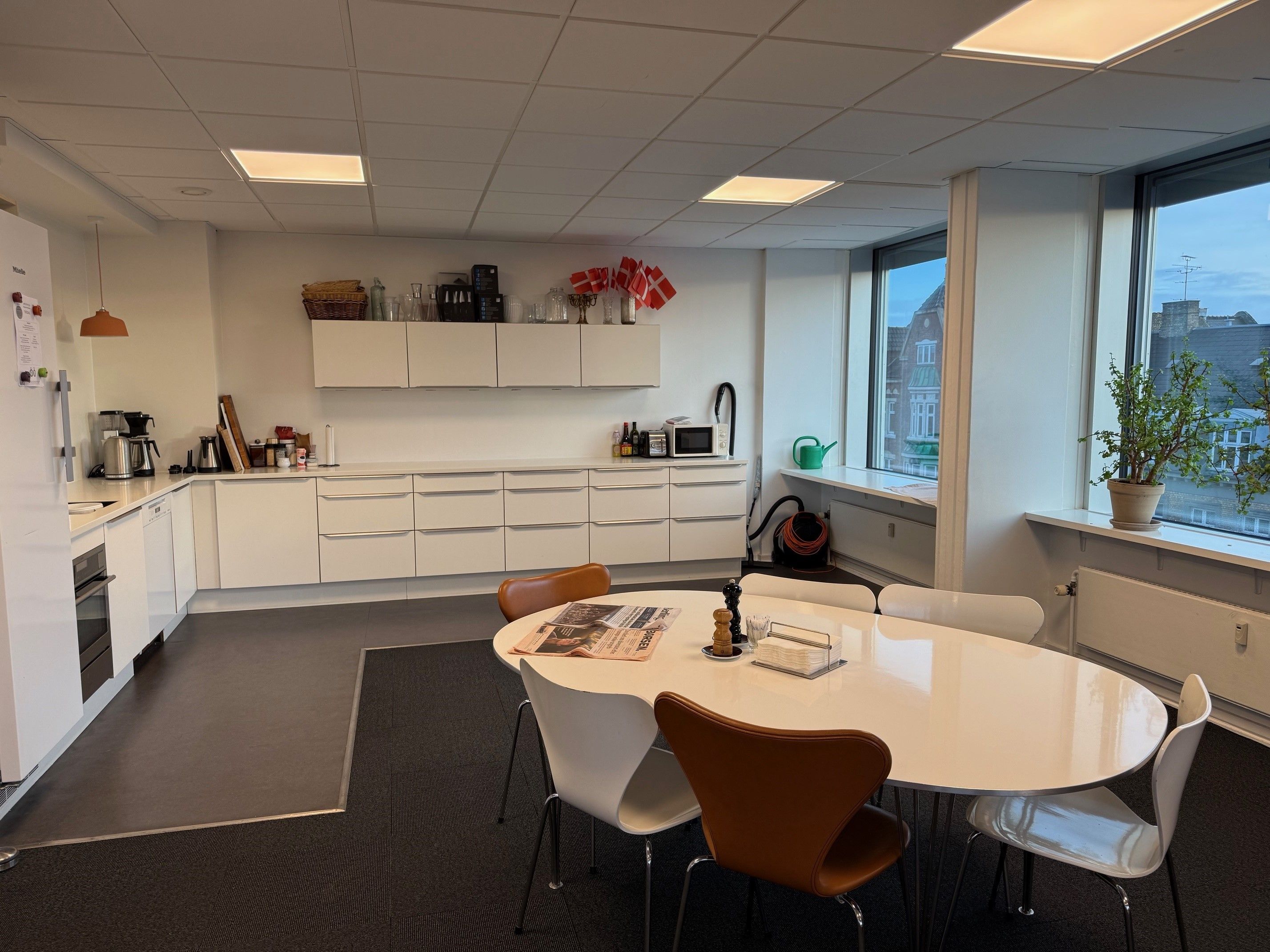 Kontorlokale med køkken, kaffemaskine, mikrobølgeovn, to bordsæt og stol, vinduer med udsigt til byen, plante i
