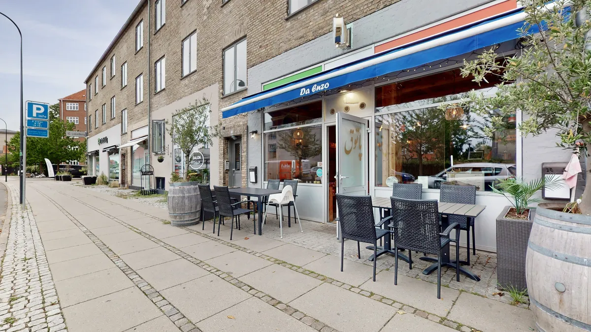 Da Enzo restaurant med terrasse og møbelsæt og bord i forgrunden og to store tønder med planter. Bygning med en stor blå