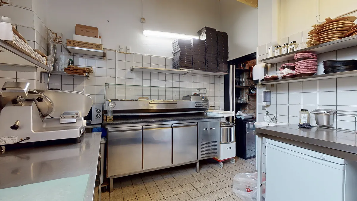 En kommerciel køkkenrum med rustfri køleskab, automatisk kødskærer og ovn samt trækasser på hylder