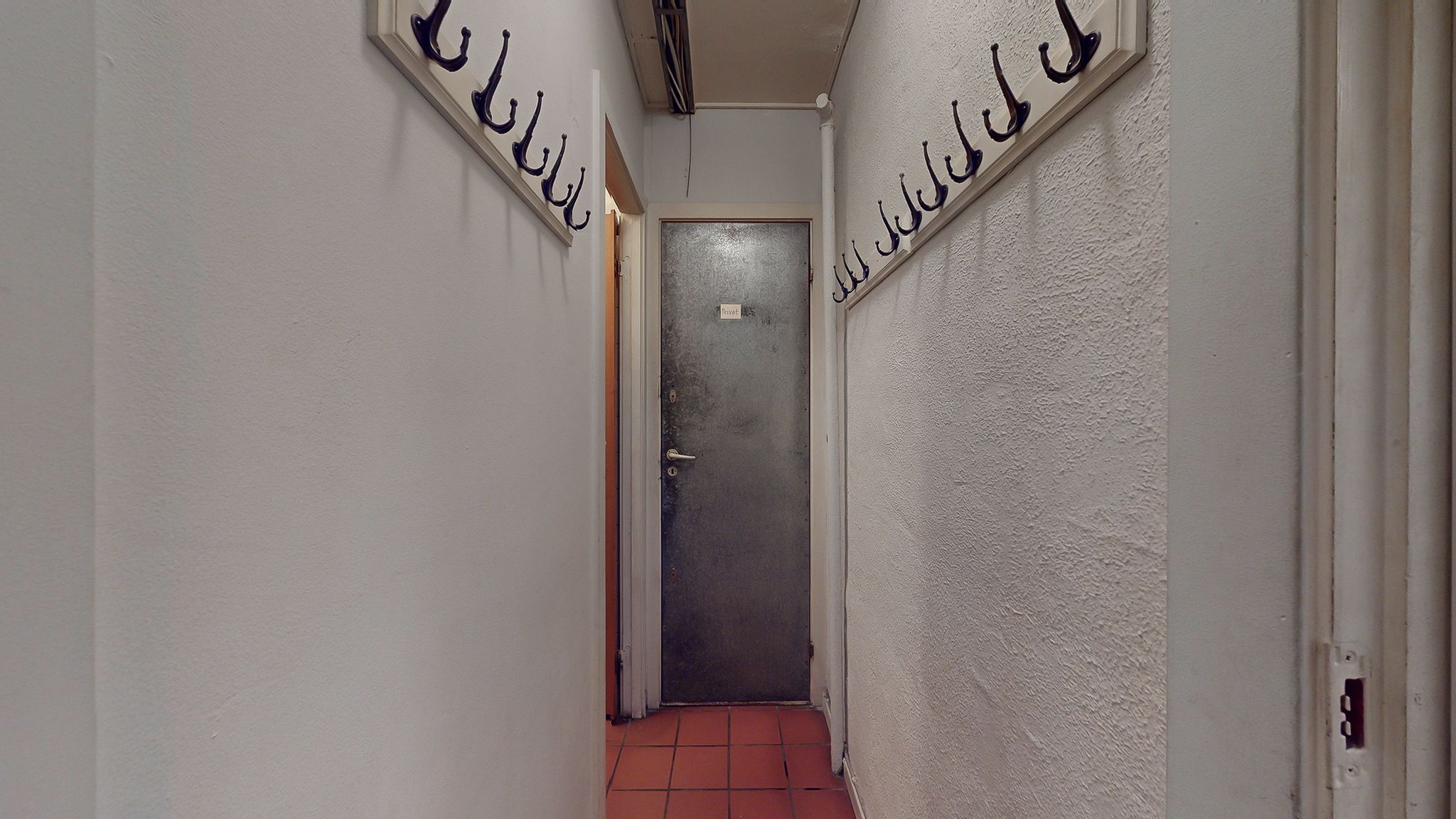 En tom gang mellem to hvide vægge med hængekroge og en åben dør i enden af gangen.