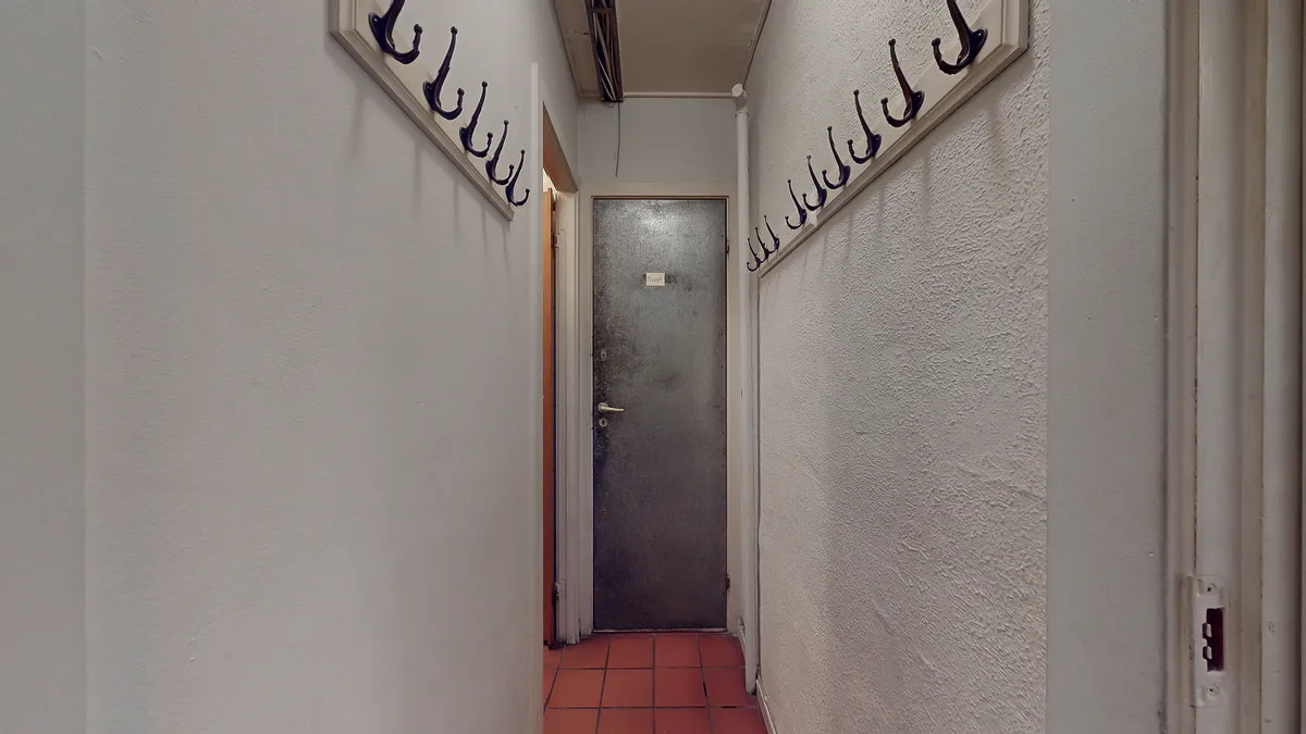 En tom gang mellem to hvide vægge med hængekroge og en åben dør i enden af gangen.