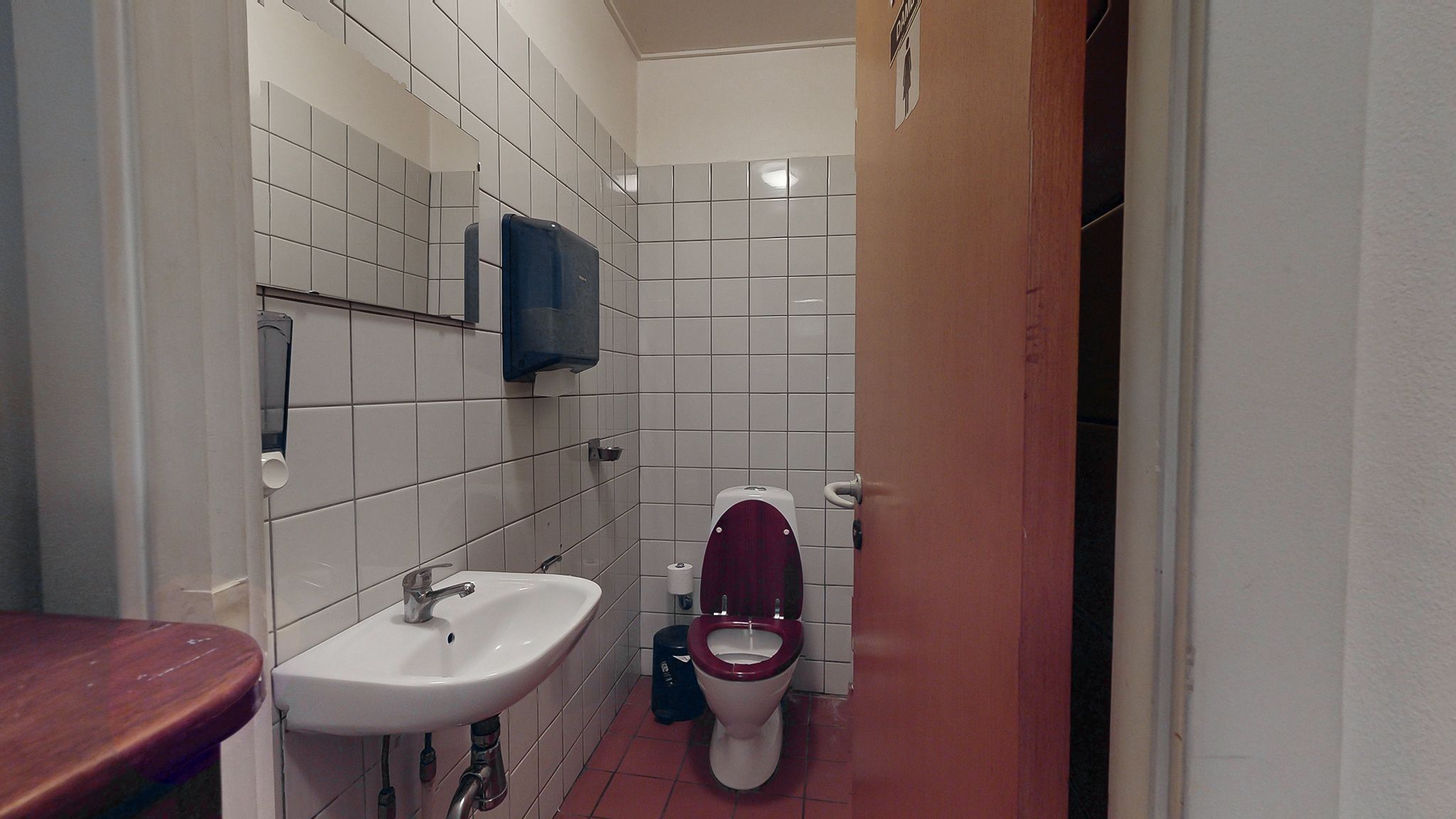 En toilet i et offentligt badeværelse med en toilet, håndvask og et håndklædeautomat samt et skel mellem rummene