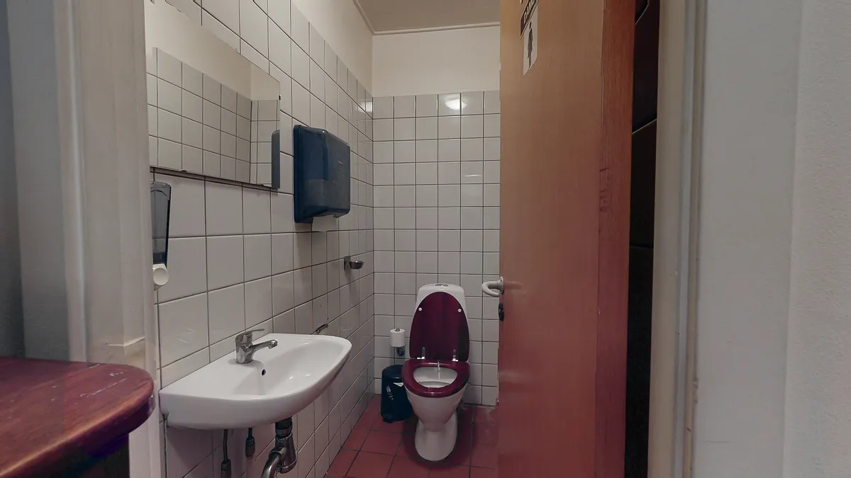 En toilet i et offentligt badeværelse med en toilet, håndvask og et håndklædeautomat samt et skel mellem rummene