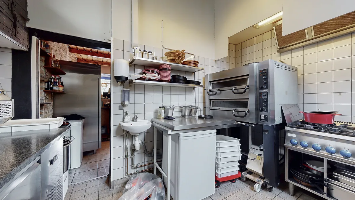 Professionelt indrettet køkken med gaskomfur, ovn, skabe og håndvask samt køleskab og flaskeholder. Køkken med hvidt flisede
