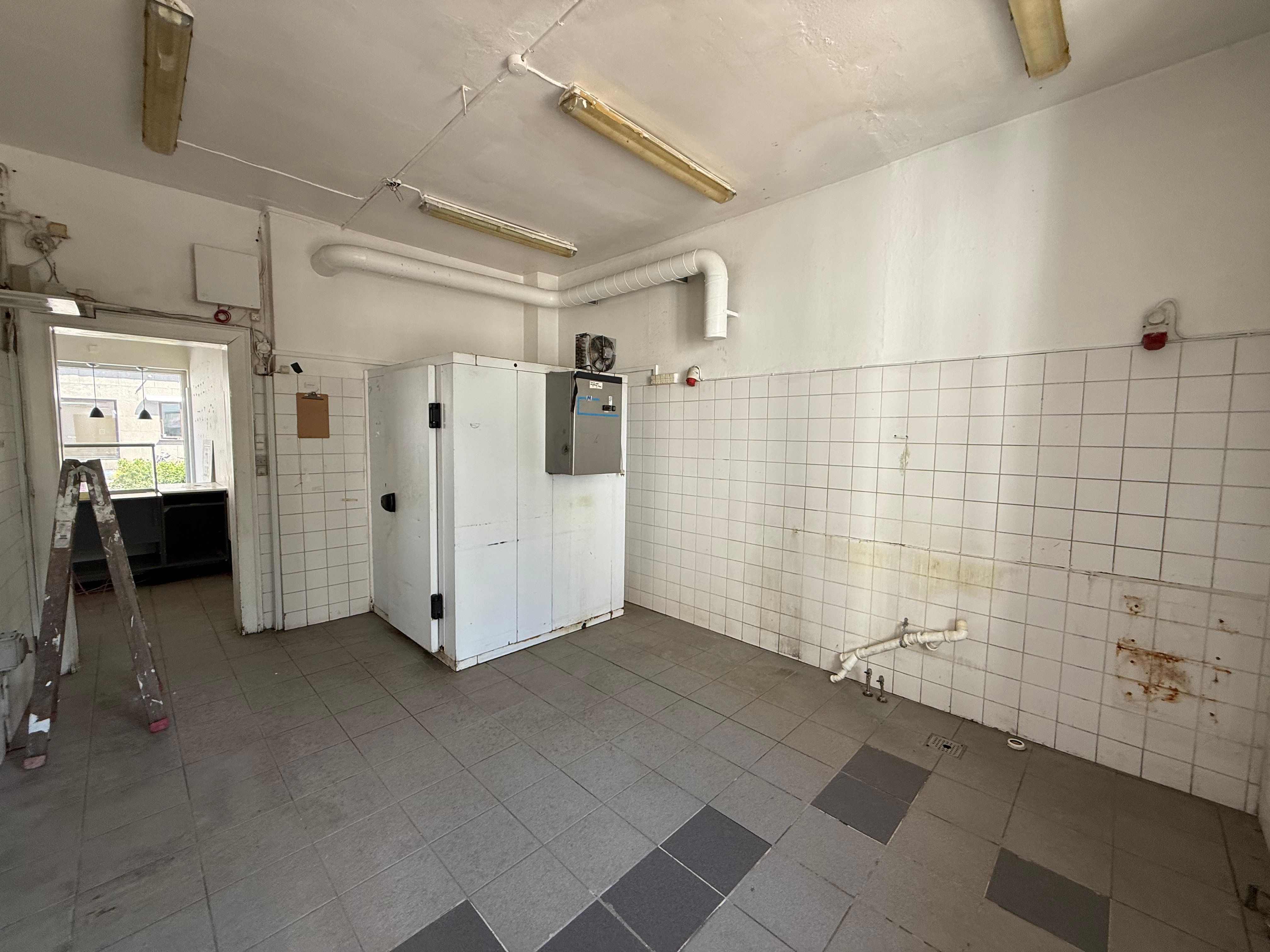 Et tomt køkken med hvide kakel og rør, gulvfliser, hængende lysekroner, luftkøler, skraldespand og stigerør