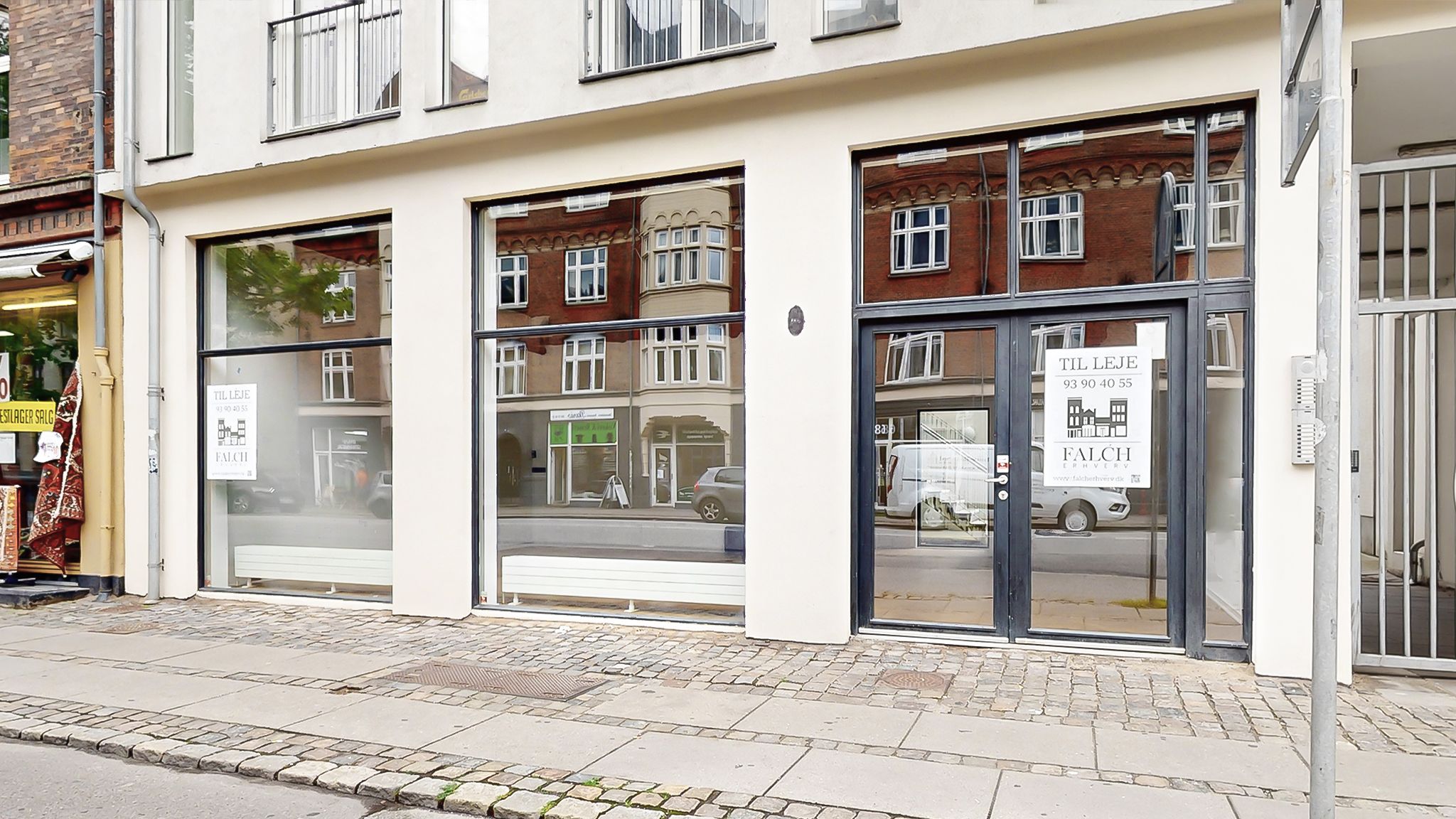 Hjørnebygning med store vinduer og glasdøre, vinduer med hvide rammer og teksten "Til Leje" på højre side