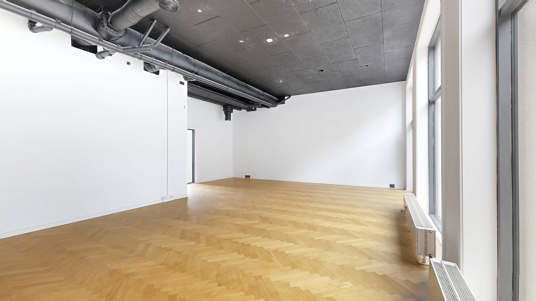 En stor tom værelse med lyse vægge og trægulv. Der er to radiatorer og røre i loftet.