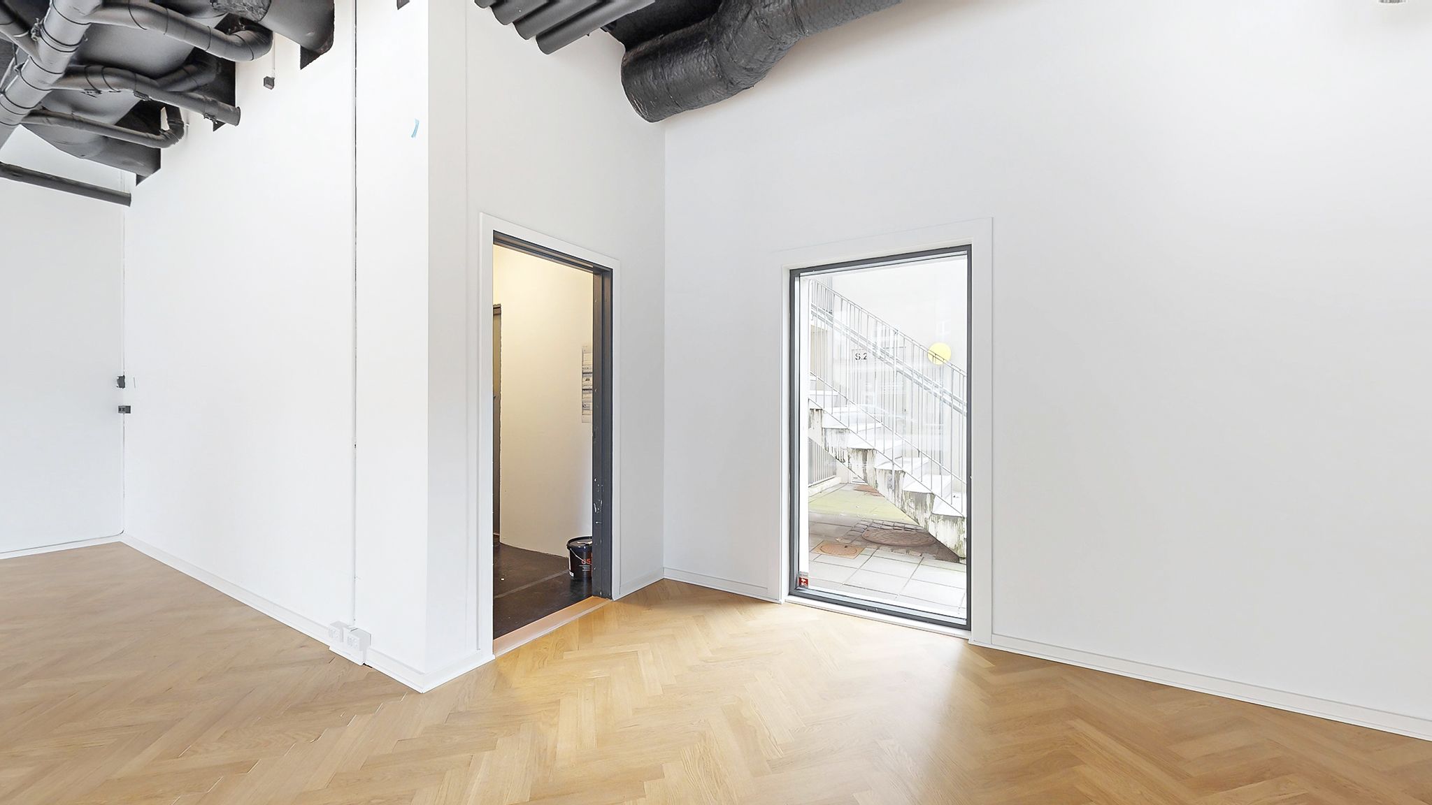 Et rum med lyse vægge og trægulv samt to døre, hvoraf den ene er åben og viser trappe til udgang