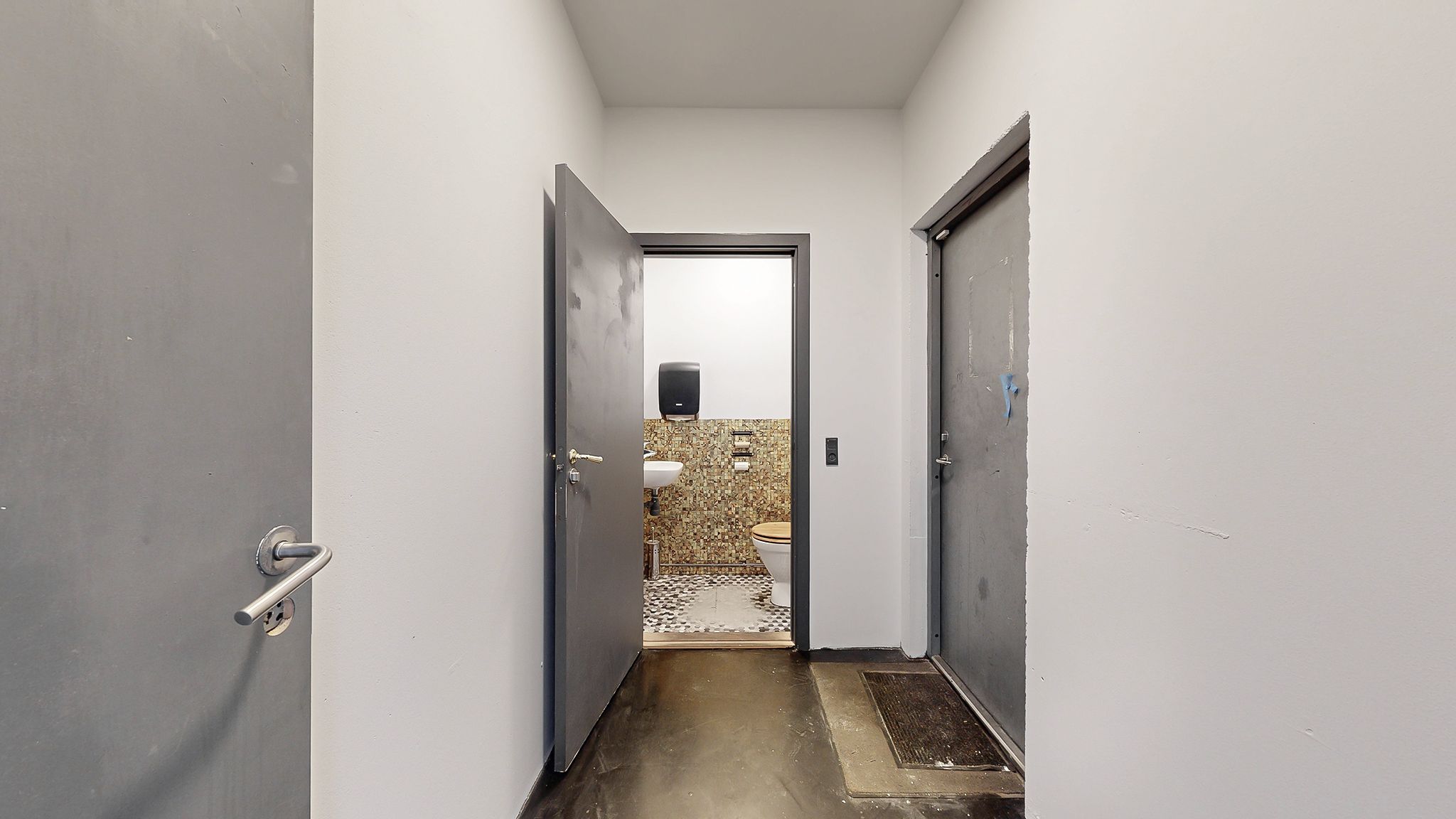 En åben toiletdør og en hængende håndklædeholder med håndklæde. Toiletrummet er lille med et toilet, håndvask og en