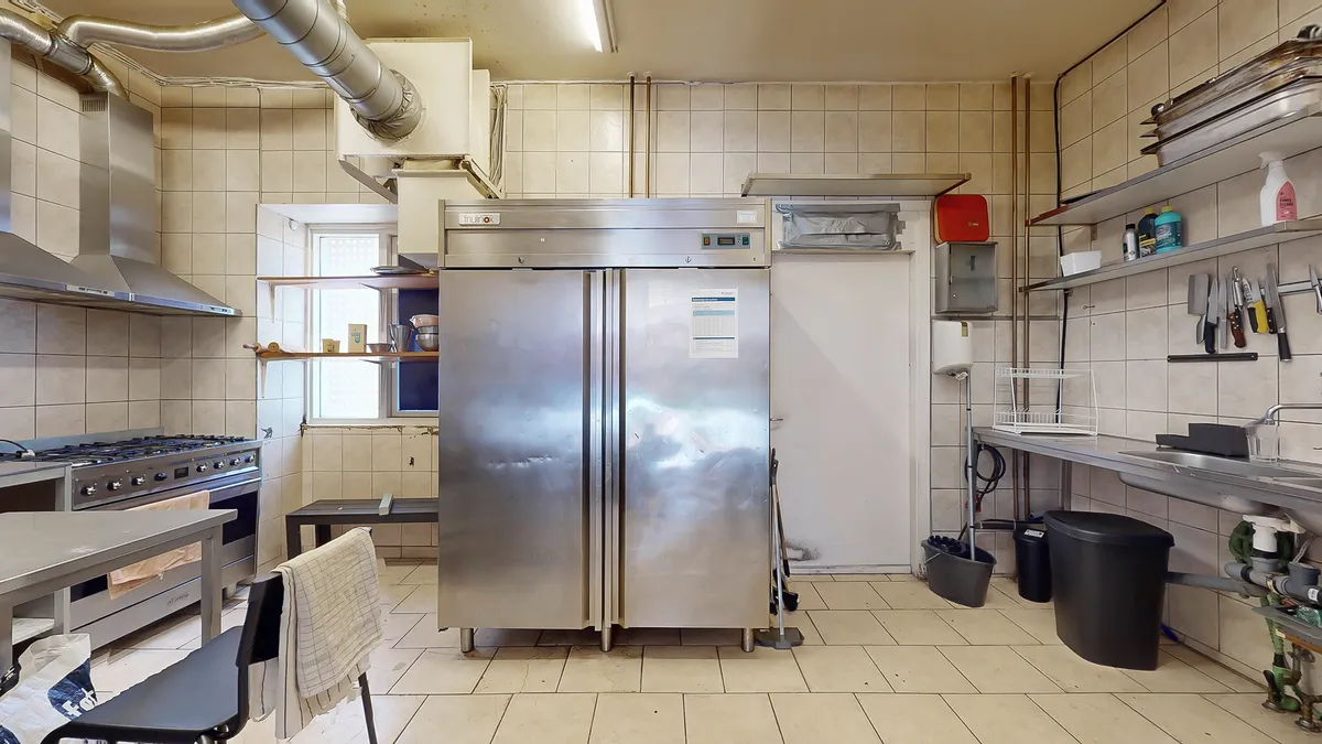 Et stort køleskab står i et kommercielt køkken med et gaskomfur, en ovn og skraldspande, hygiejneprodukter og knive på