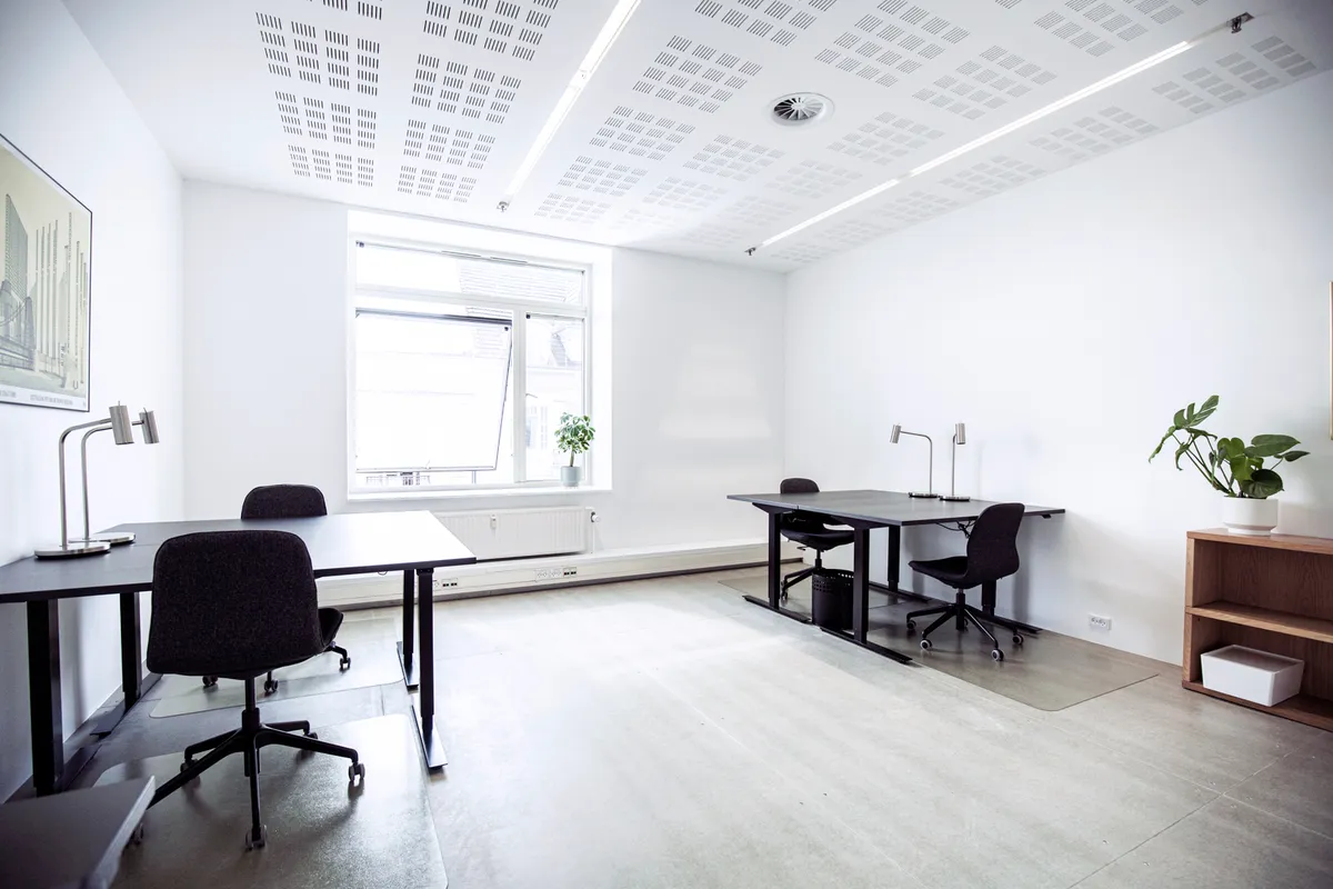 Kontorfællesskabet med minimalistisk indretning, to bordsæt med stole, plante i hjørne og vinduesudsigt over byen