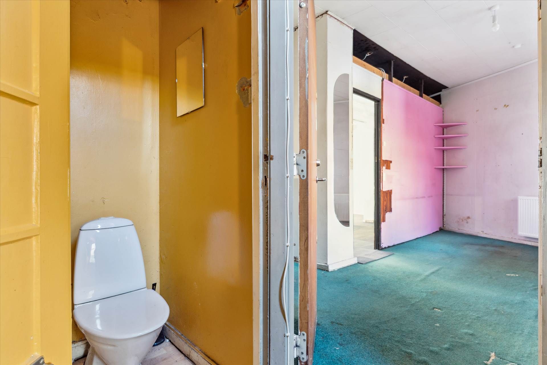 En gammel to-etagers lejlighed med en toilet i en gul væg og en åben dør til et tomt rum med lysegul og rosa væg