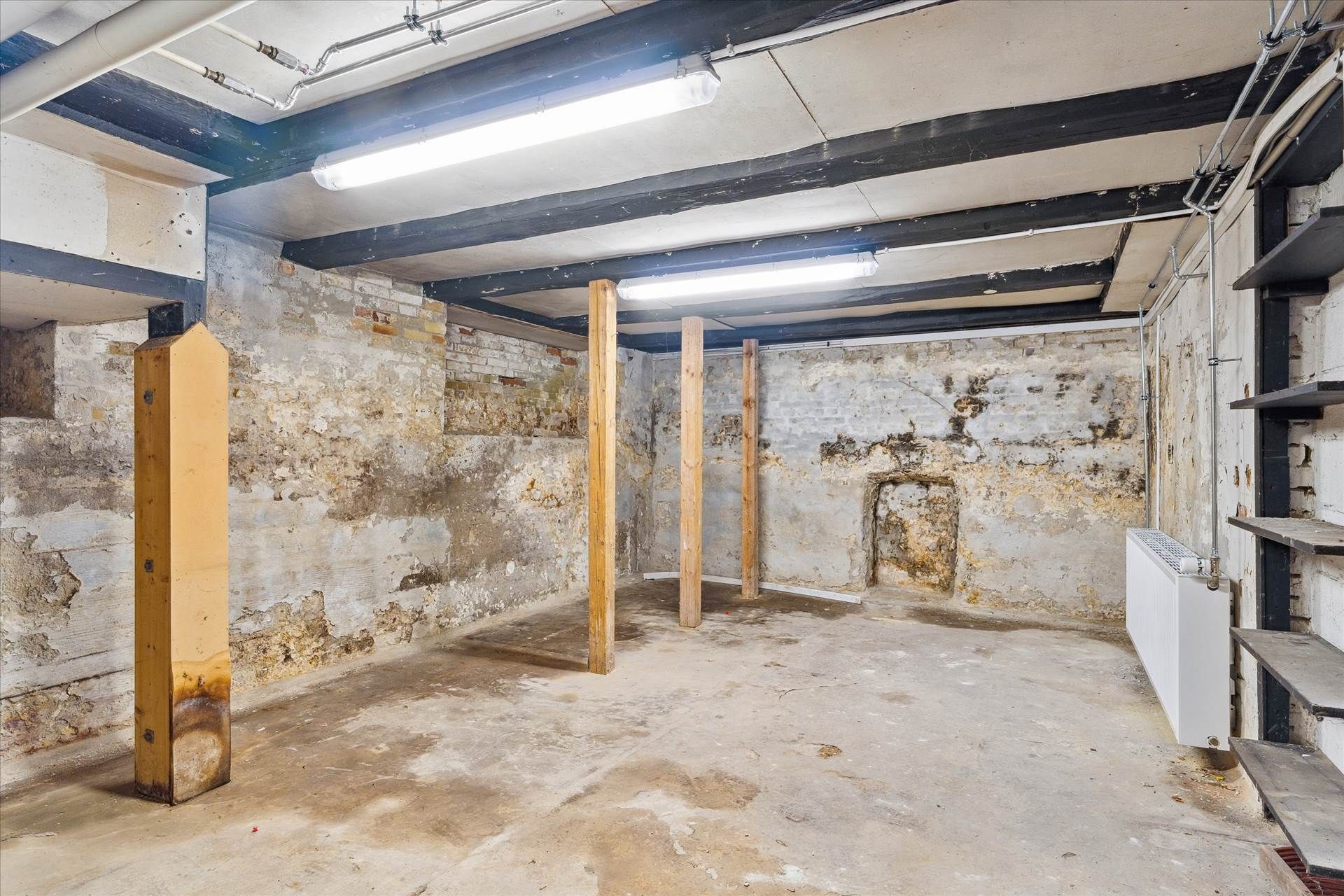 Et gammelt og forfaldent lagerrum med fugt og skimmel på væggene, lysekroner og gulv samt stående bjælker og bjælkeafstande