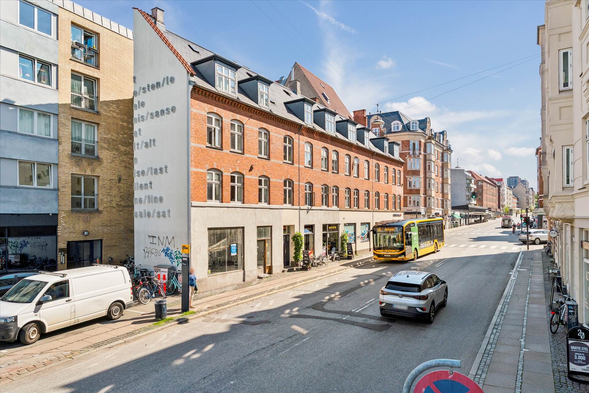 En bus kører gennem gaden, hvor der er parkerede biler og cykler langs fortovet med butikker og graffiti på bygningerne