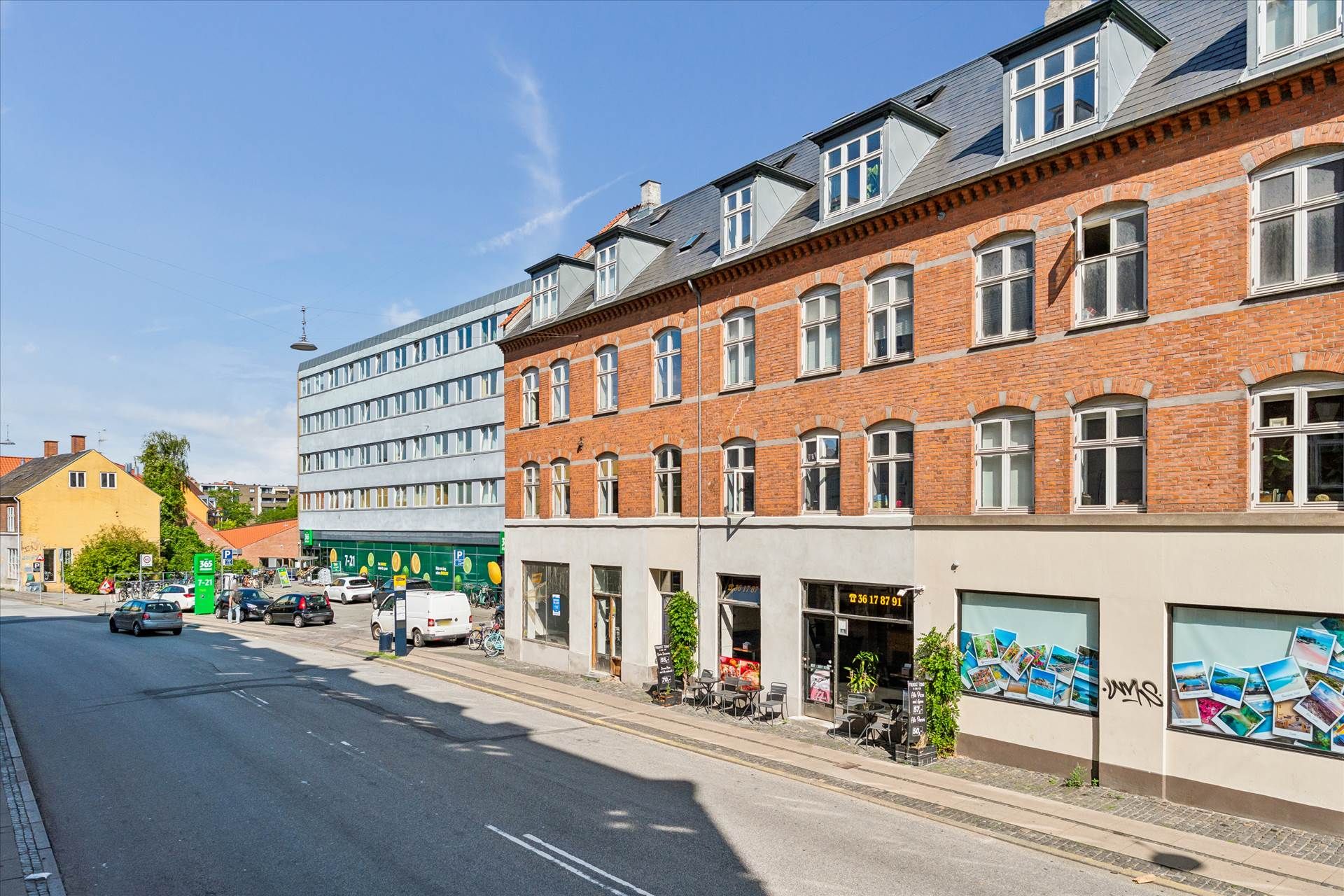En bred gågade med parkerede biler og grønne planter foran et rødt lejlighedsbyggeri og et stort byggeprojekt