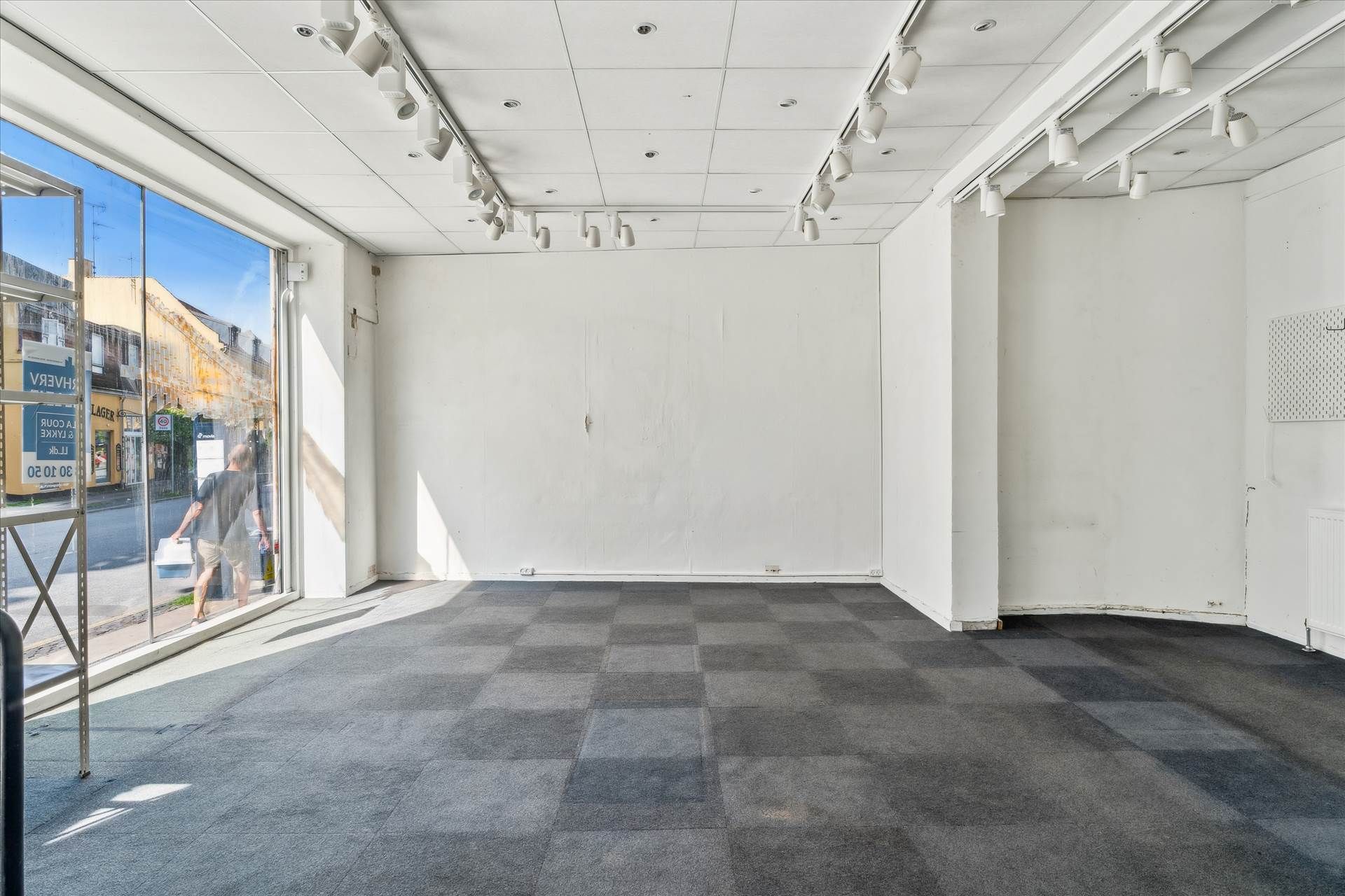 Et ubeboet erhvervslejemål med store vinduer og lyse loftsspots er til leje. Der er karnapper og gulvbelægning med karreer.