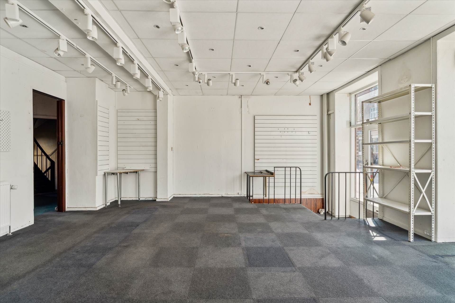 Et stort, tomt lokale med lysegule vægge, hvidt loft, carpettering, metalbøjler og høje vinduer, der giver godt lys