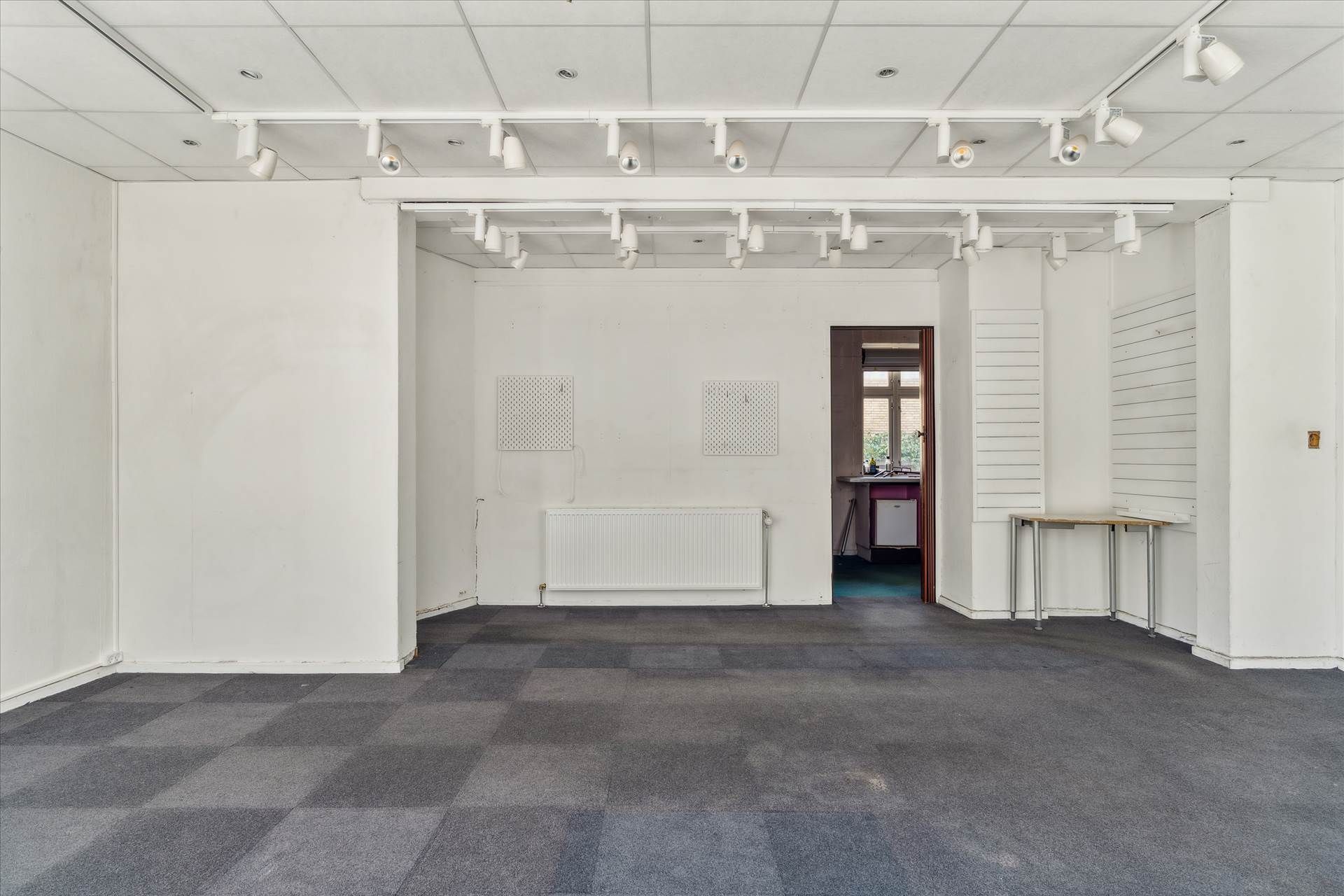 Et tomt rum med lyse loftslampe, hvid væg og gulv, to møbler og et skur uden for døren