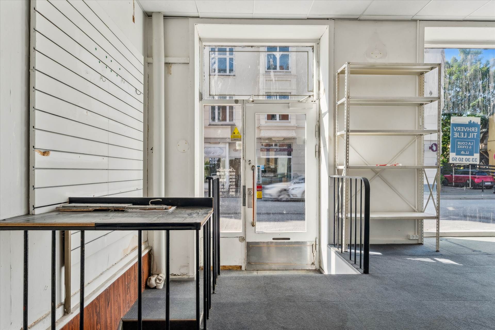 Tømt erhvervslejemål med glasdør og vinduer på Valby Langgade, tæt ved en bilparkeringsplads med biler og en terrasse med
