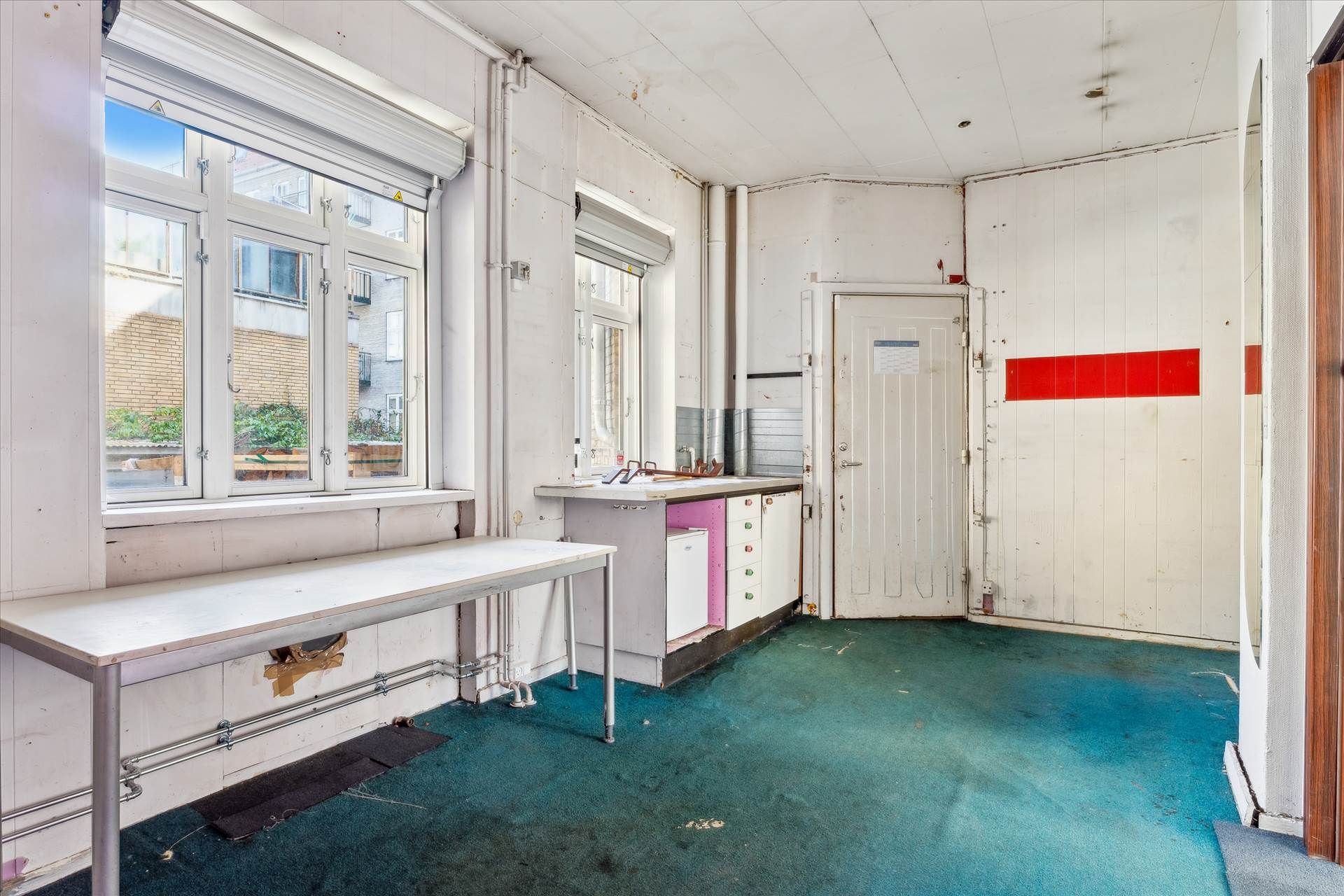 En tomt og ubenyttet erhvervslokal med en stor åben dør, et langt køkkenbord og en gulvbelægning i blåt tæppe