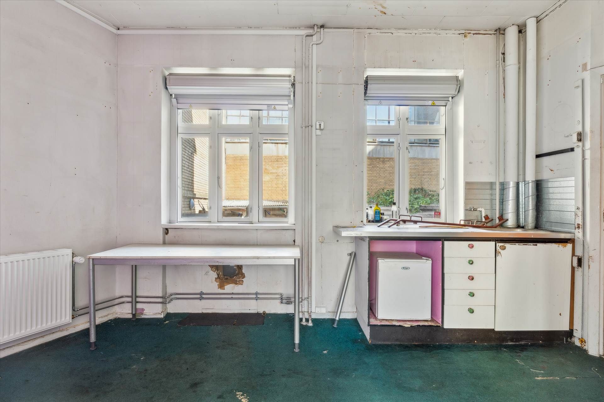 En ubenyttet værkstedslokal med gulgulvt og to vinduer, hvor der er et skarpt køleskab og en ståbord samt et radiator og rør