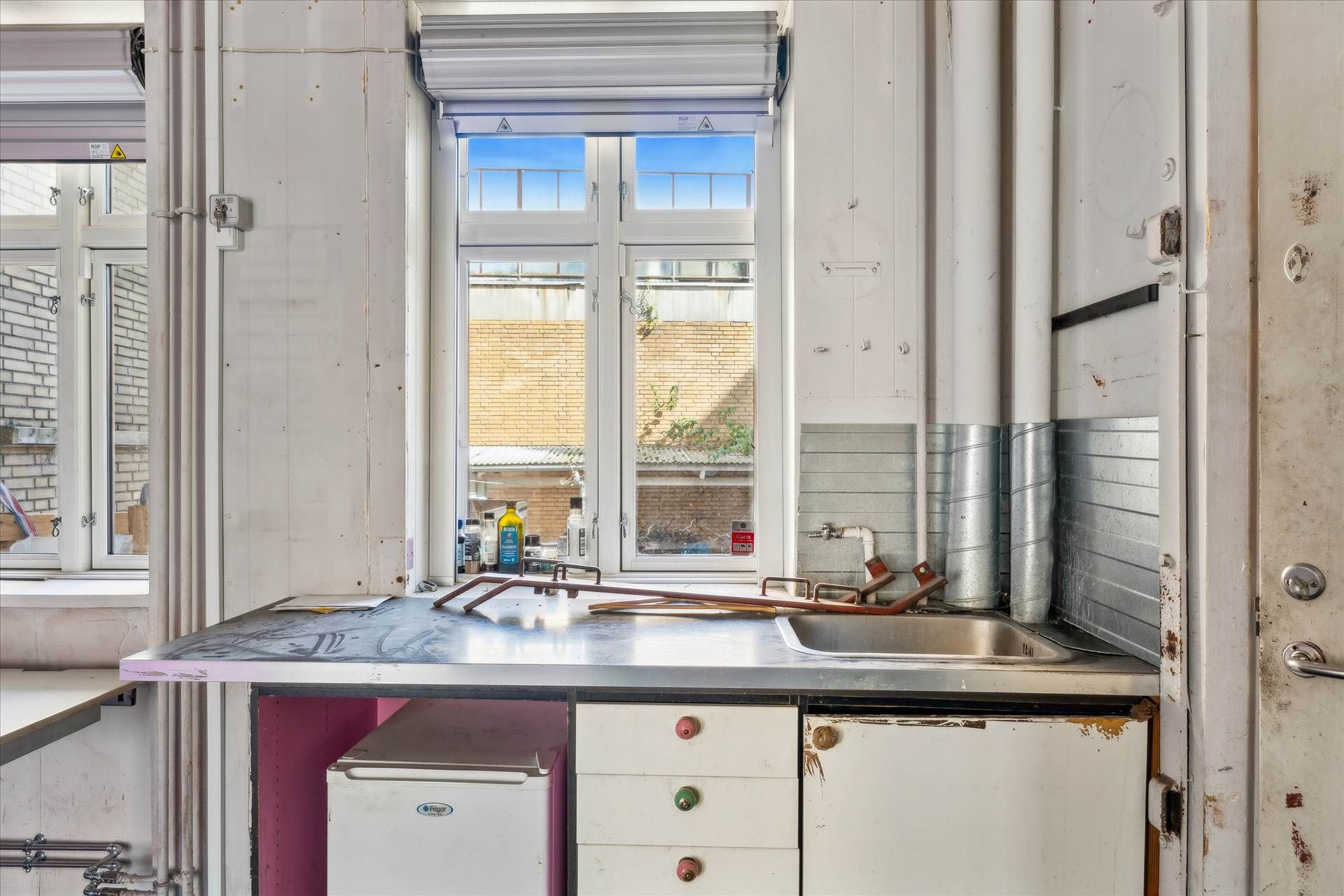 En enkelt køkkendel med skrivebord og en stor skraldespand, en rusten ovn samt en vandhane og et ældre køleskab i køkkenets