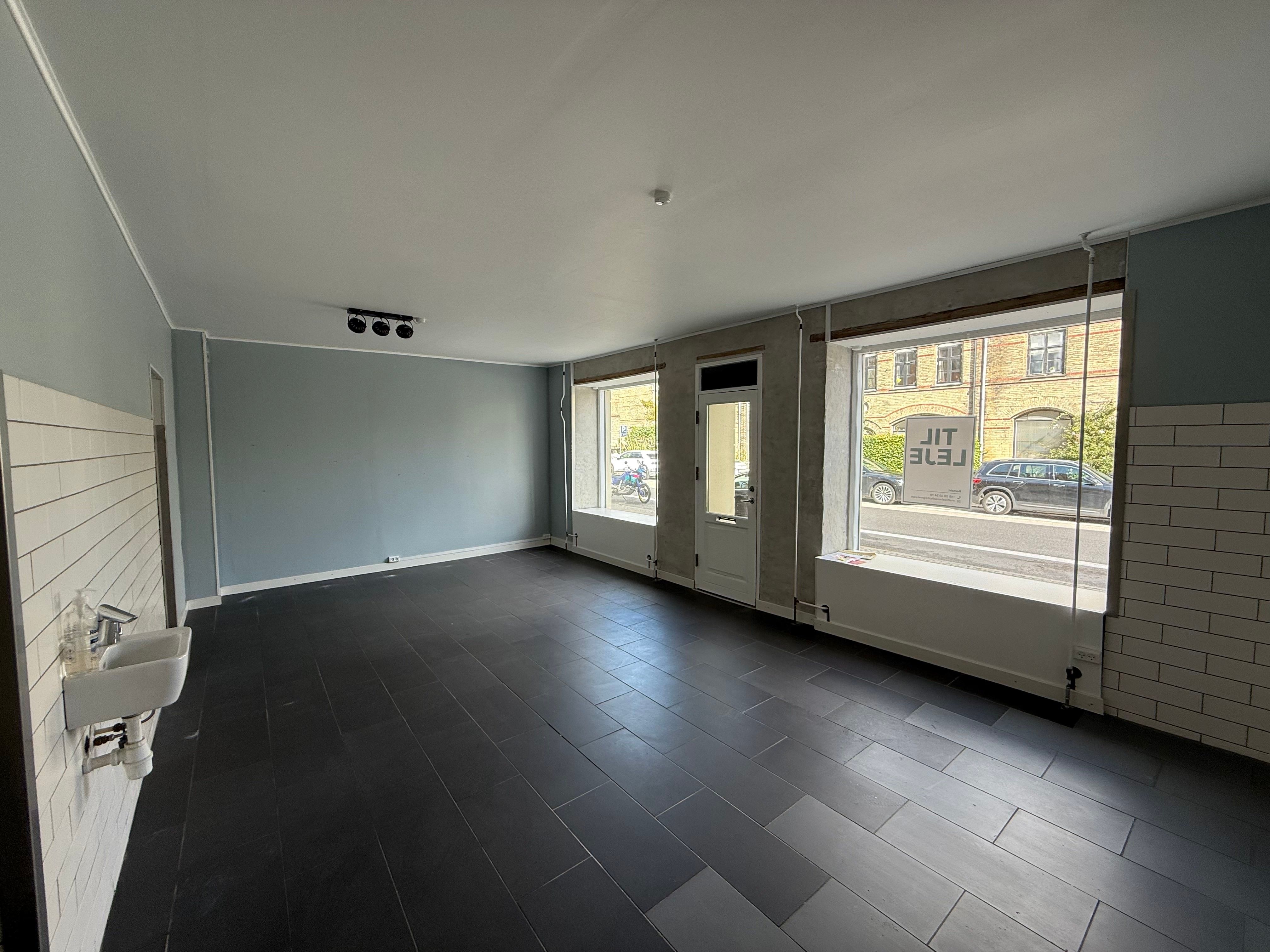 Tom rum med højt loft, hvide vægge og sort gulv samt to store vinduer med udsigt til gaden og en bilparkeret ved en rødt hus