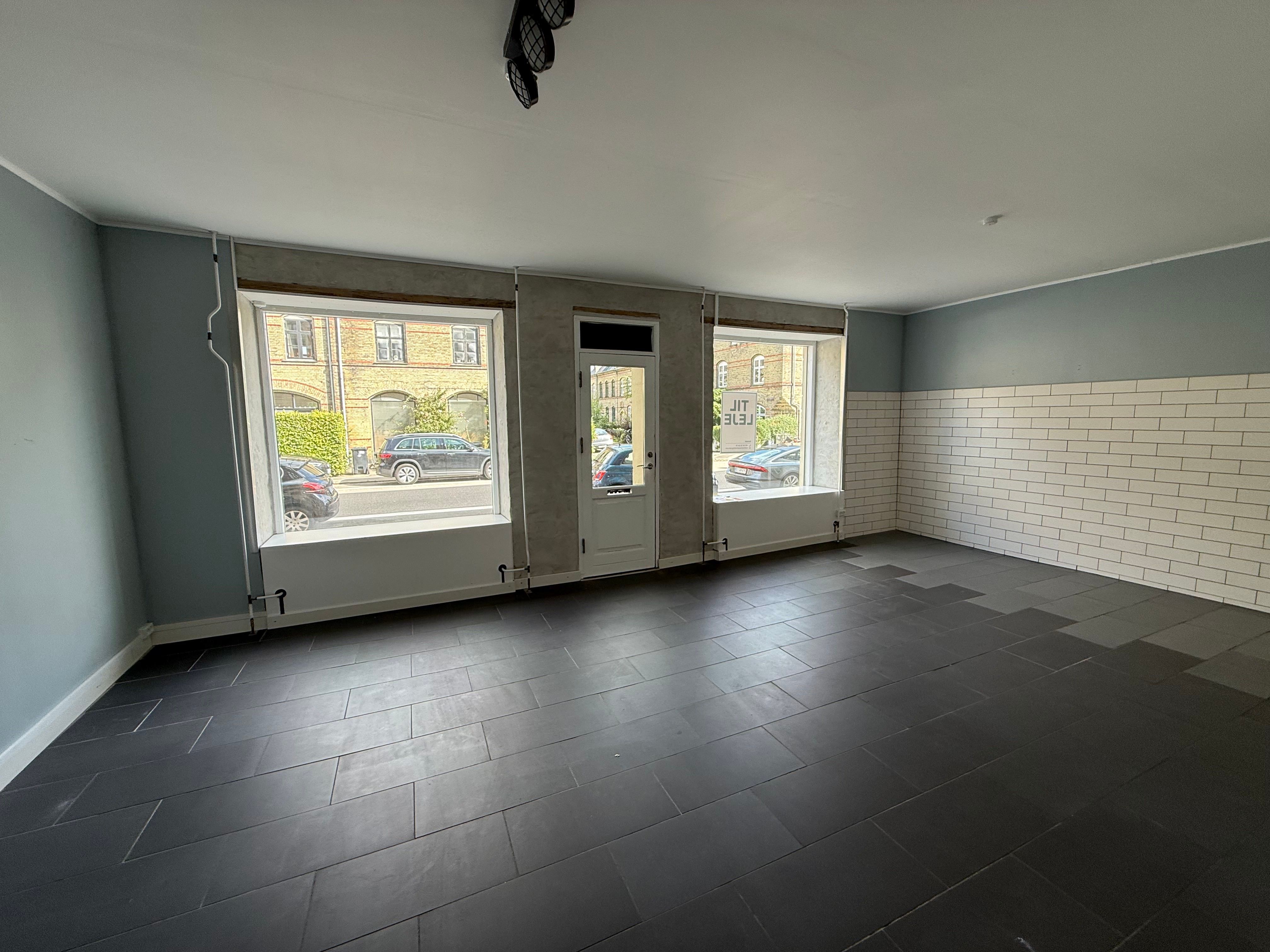 Et stort, tomt lokale med vinduer, hvor der står "Leje" på én af dørene, og hvor der er hvide mursten og en lysegrå væg