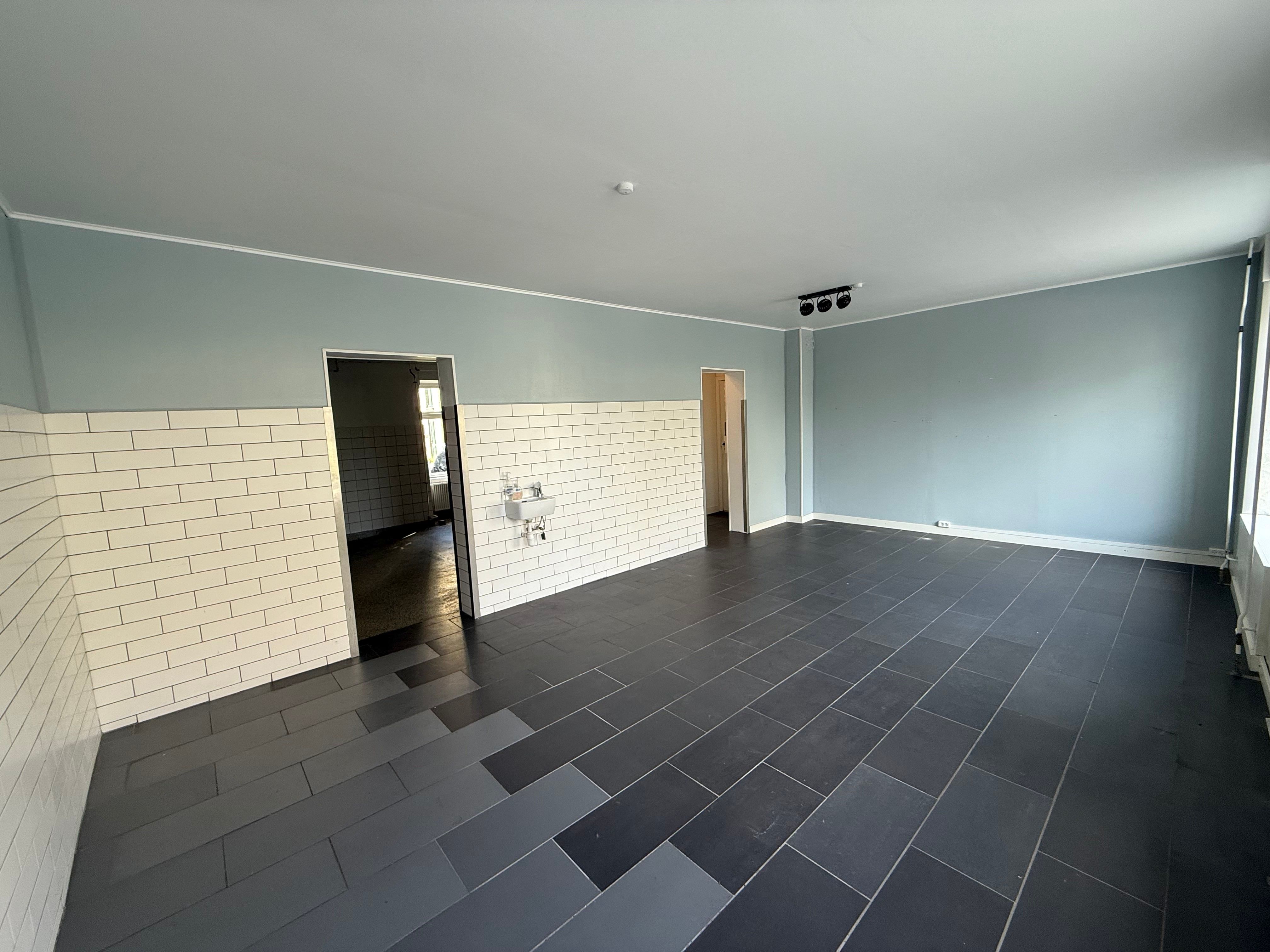 En stor, ren og rummelig butikslokal med lysegrå vægge, sort og gråt gulv og to åbne døre til andre rum.