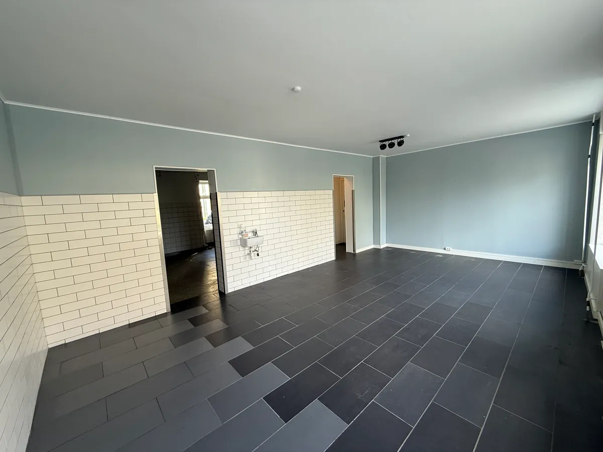 En stor, ren og rummelig butikslokal med lysegrå vægge, sort og gråt gulv og to åbne døre til andre rum.