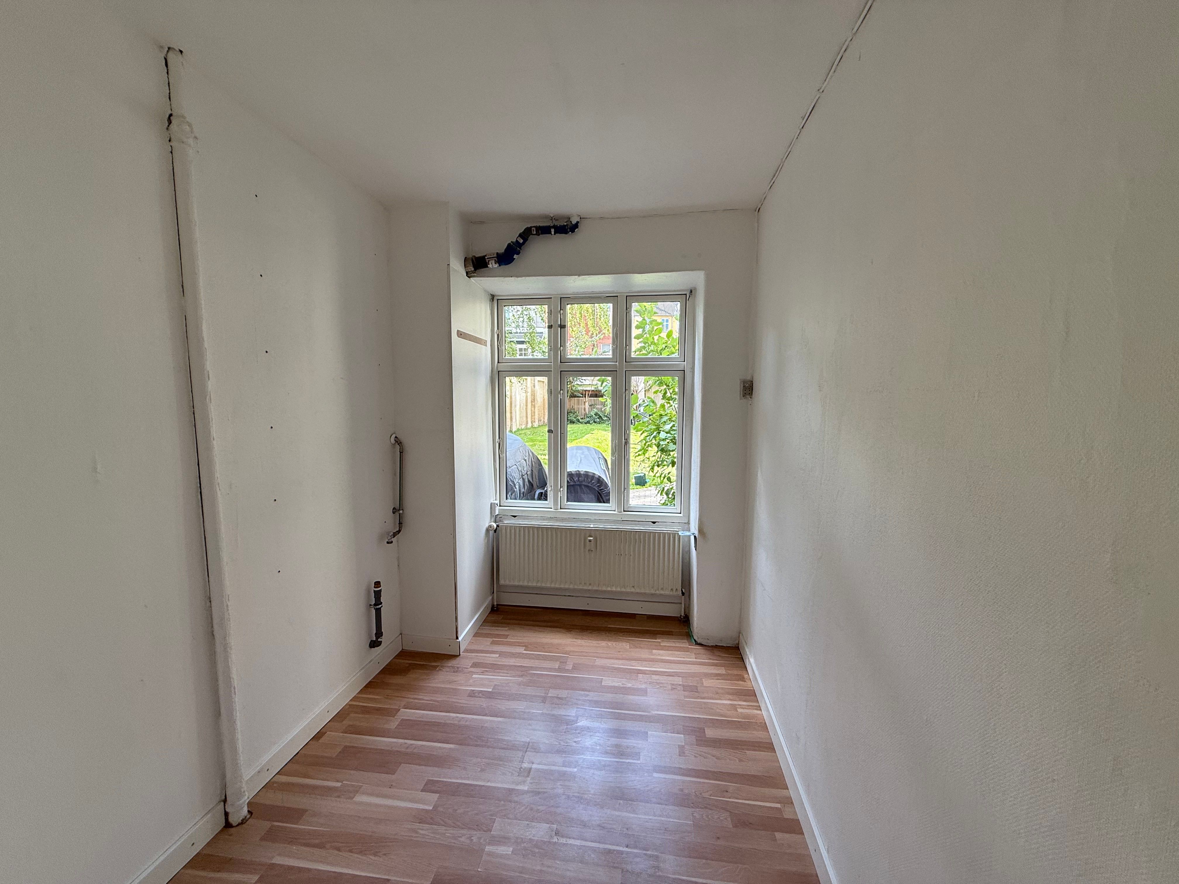 En tom gang med hårdt trægulv, hvidvaskede vægge og et vindue med udgang til haven