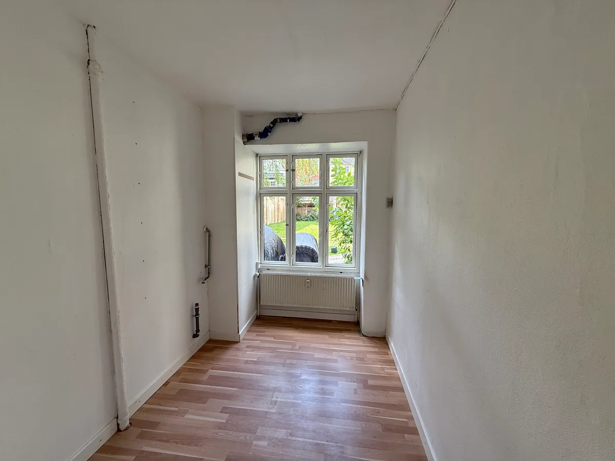 En tom gang med hårdt trægulv, hvidvaskede vægge og et vindue med udgang til haven